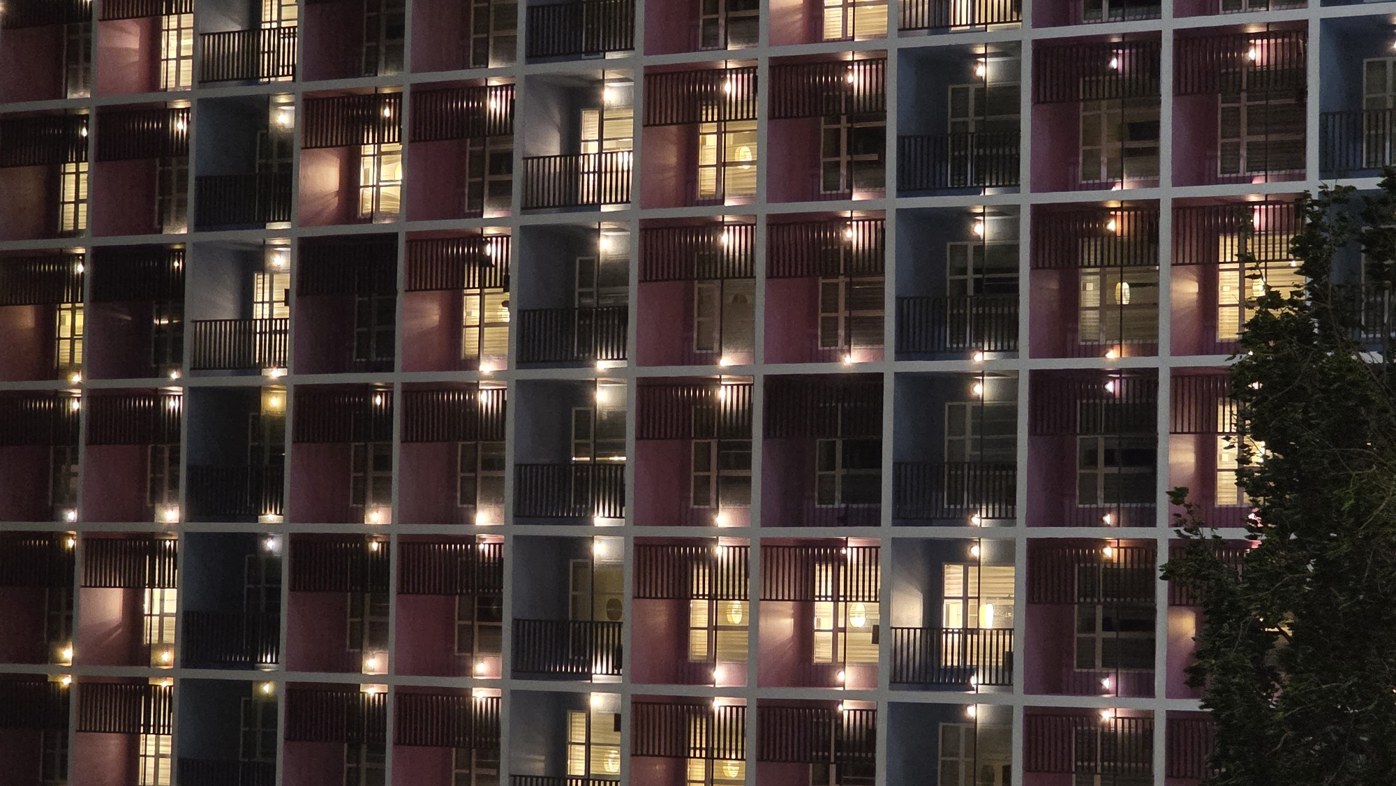 The image shows a building facade in Nha Trang, Vietnam, at night. The building is a multi-story structure with a grid-like pattern of square window openings. The wall spaces between windows are colored in alternating shades of pink and blue. Each window appears to have a small balcony with dark railings. Many of the windows are illuminated from within, suggesting that the building is occupied. The lights produce a warm, yellowish glow. On the right side of the image, a tree is partially visible, with its leaves appearing dark against the illuminated building.