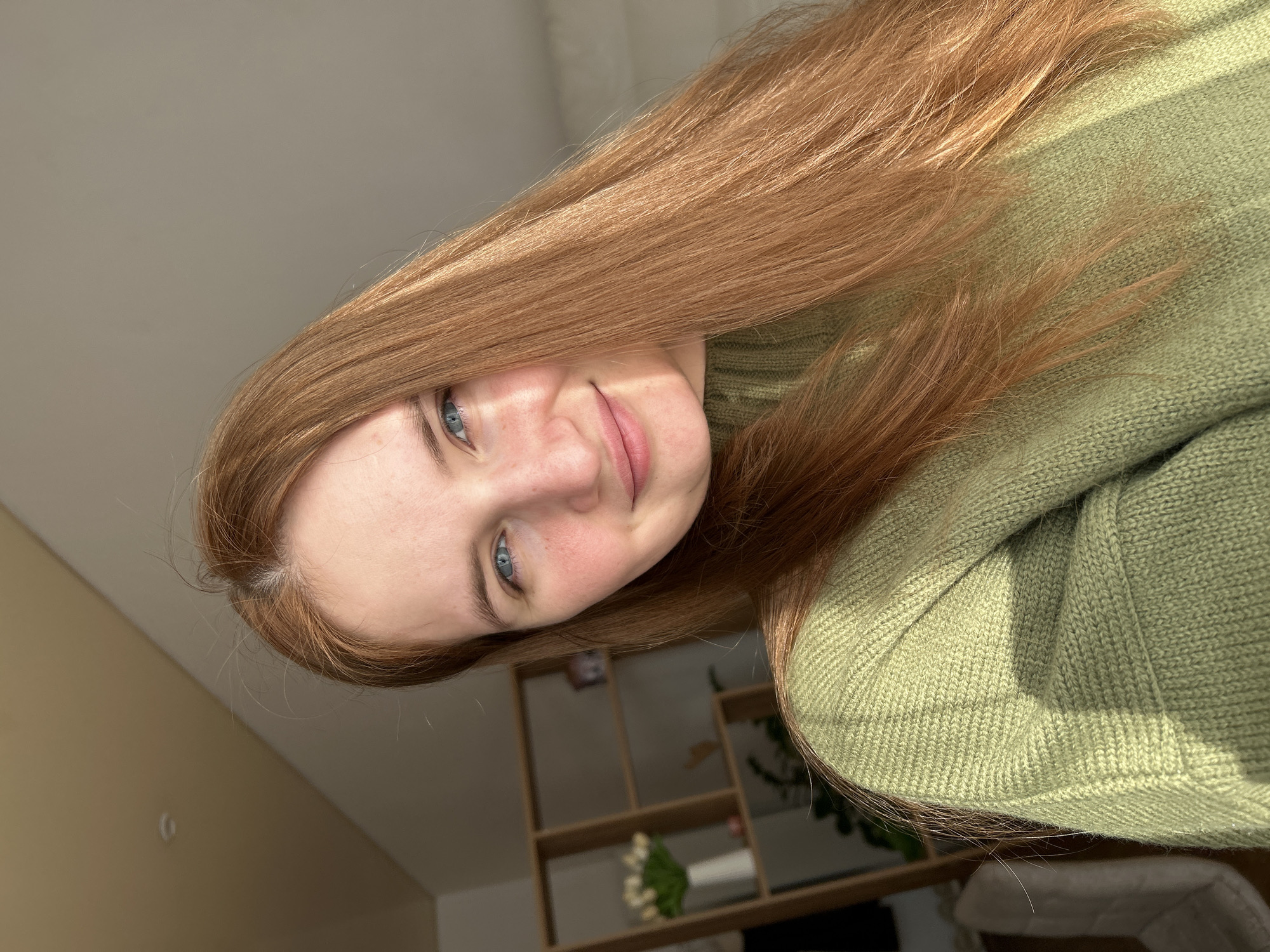 A young woman with long, reddish-brown hair and blue eyes is smiling. She is wearing a light green sweater. Behind her is a light-colored wall and a shelving unit with a vase of white flowers on one of the shelves. The image was likely taken indoors. No further details can be confirmed regarding the specific location in Chaikovsky, Russia.