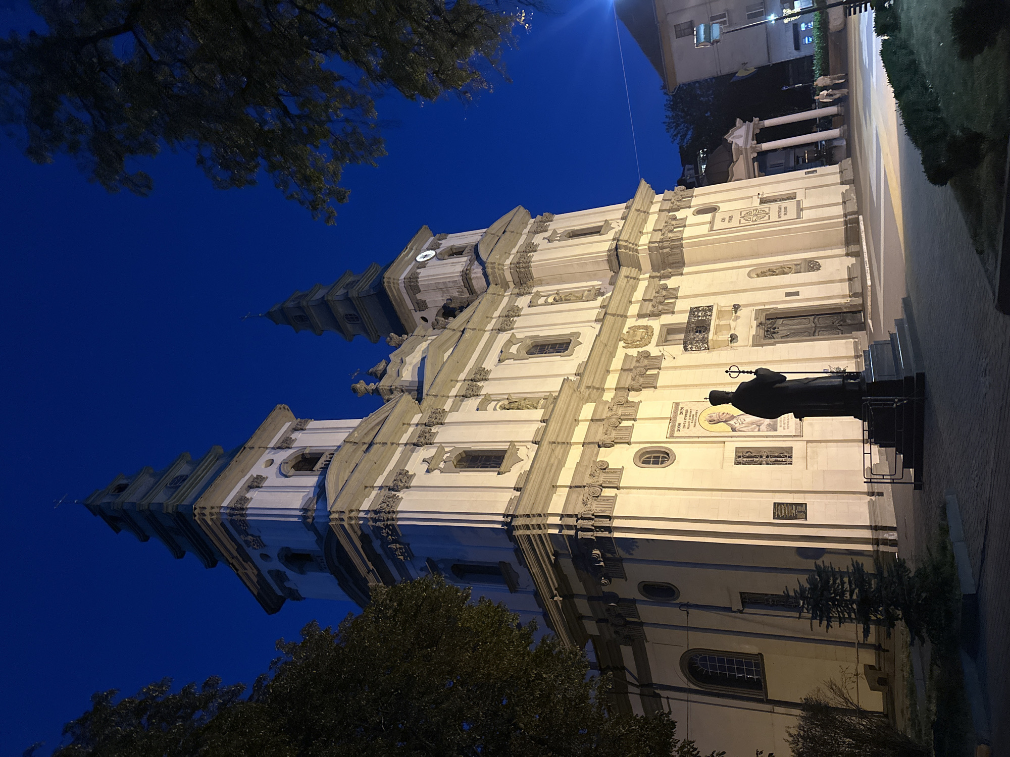 The image depicts the Ternopil Cathedral of the Immaculate Conception of the Blessed Virgin Mary in Ternopil, Ukraine, at night. The cathedral is illuminated by external lighting, highlighting its white facade and twin spires. In the foreground, a dark silhouette of a statue of a robed figure holding a staff stands on a pedestal, facing the cathedral. The sky is a deep blue, indicating twilight or night. Trees with dark foliage frame the top and left sides of the image. Paved pathways and some landscaping are visible in front of the cathedral. In the background, another building with a colonnade and a lamppost with a modern light fixture are present. Two individuals are walking in the distance near the colonnaded building. The overall scene is one of solemnity and architectural grandeur.