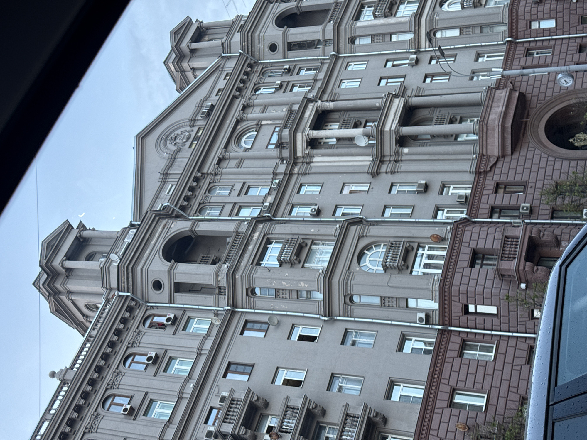 The image shows a tall building with a gray facade, adorned with architectural details like arched windows, columns, and balconies. Numerous windows, some with air conditioning units attached, are visible. The building appears to be located in Moscow, Russia. A section of the building's facade is brick-like in texture and brownish-purple in color, with a large circular window-like structure. A portion of a car window is visible on the left and right edges of the image, implying that the picture was taken from inside a vehicle. The sky above the building is overcast.
