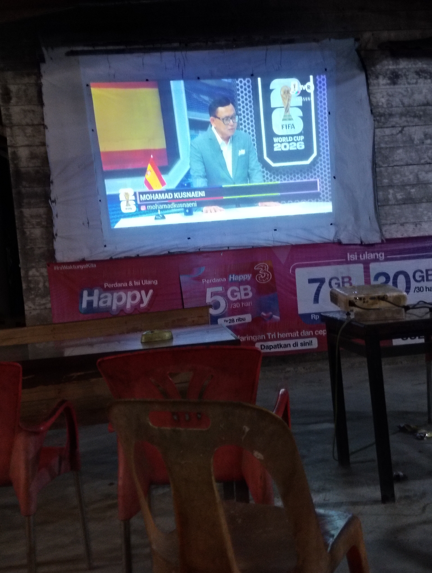 The image depicts an outdoor projection screen displaying a broadcast related to the FIFA World Cup 2026, featuring a commentator named Mohamad Kusnaeni. In the foreground, several red and brown plastic chairs are visible, suggesting a viewing area. Behind the screen, a banner for "Happy" mobile data plans is displayed, indicating a commercial establishment. The scene appears to be in an urban or semi-urban environment, possibly a street-side eatery or gathering spot in Payagaboh, Indonesia. The lighting suggests it is evening or night.