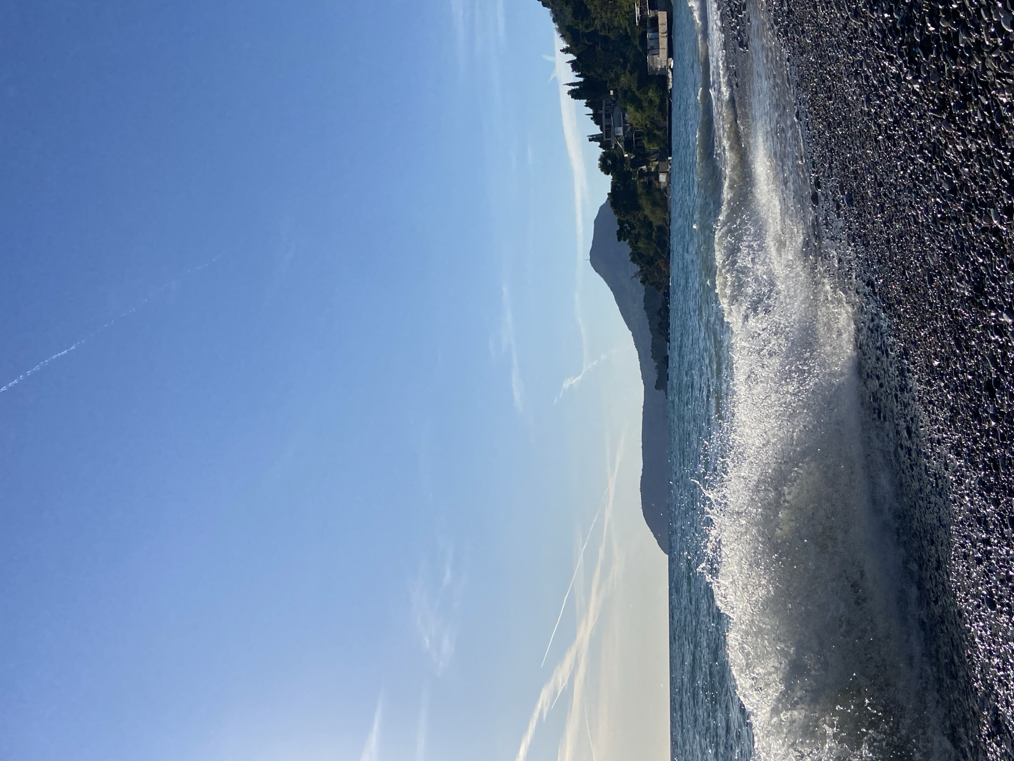 The image captures a coastal scene near Bar, Montenegro, dominated by a bright blue sky streaked with airplane contrails. To the right, dark, wet, glistening pebbles compose the beach, where a wave crashes violently, creating a splash. In the background, a mountain rises beyond the coastline, partly obscured by a lighter haze. A line of trees and buildings is visible at the top of the hill.