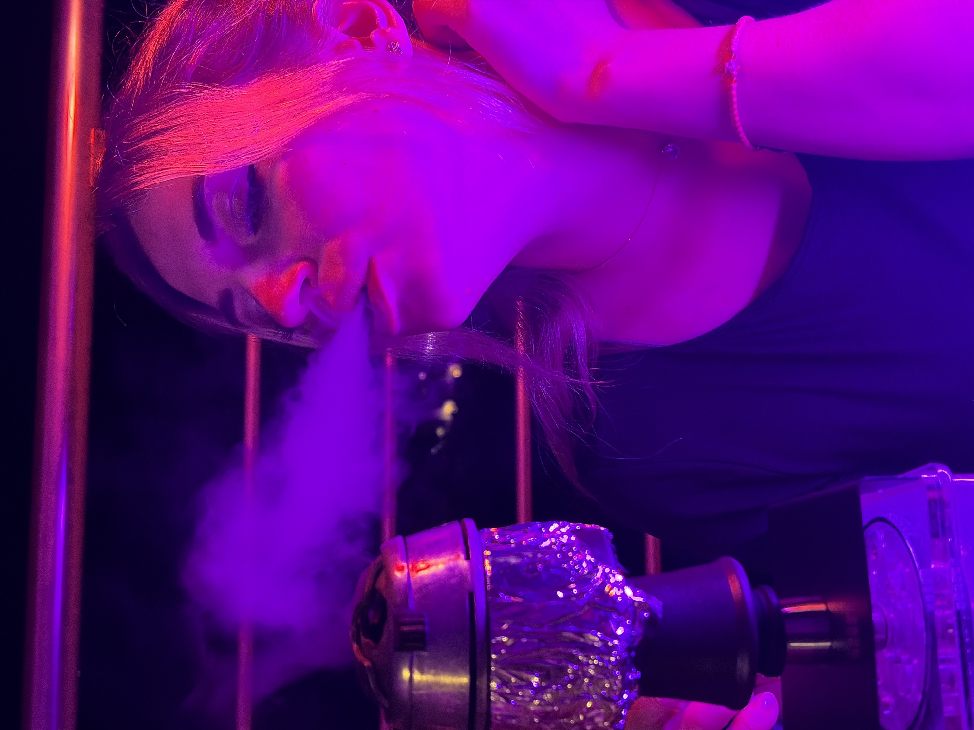 A woman is captured in a medium shot, exhaling smoke from a hookah. The scene is dimly lit with vibrant purple and pink lighting, creating a moody atmosphere. The woman's face is partially illuminated, with her eyes closed as she exhales. A hookah pipe is visible on the left side of the frame, its metallic bowl covered in foil, with a plume of purple smoke emanating from it. The background is dark, with some distant lights suggesting an urban setting, possibly the city of Budva, Montenegro. The woman is wearing a black top and a delicate necklace. Her hand is raised to her neck, with a thin bracelet on her wrist. The overall impression is one of a social setting, possibly a lounge or bar, with the focus on the act of smoking the hookah.