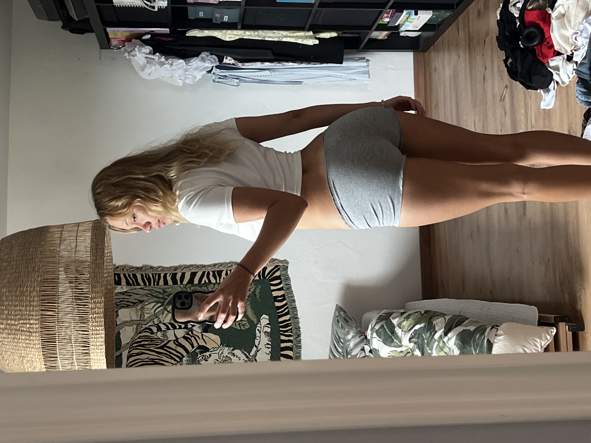 A young woman with long blonde hair is taking a mirror selfie. She is wearing a white t-shirt and grey ribbed shorts. She is standing in what appears to be a bedroom or dressing area. Behind her, a shelf unit is filled with books and other items. To her left, a large woven basket is visible, and to her right, a bed with a patterned duvet and pillows can be seen. The floor is made of wood planks. The woman's expression is neutral as she looks into the camera. The lighting suggests it is daytime. The location is Mühldorf a. Inn, Germany. There is no visible text in the image.