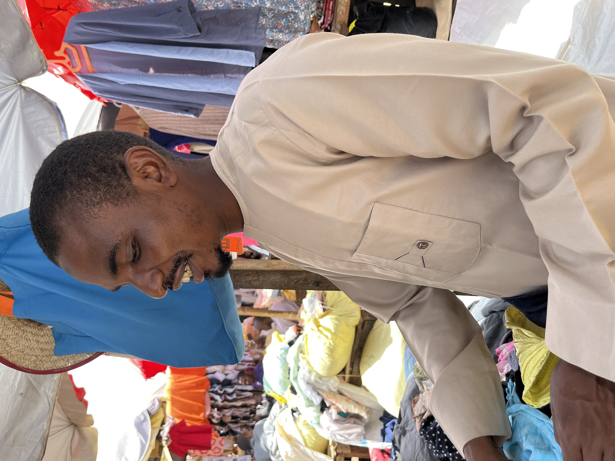 An eye-level indoor shot, rotated 90 degrees counterclockwise, shows a man in a tan shirt standing behind a table in a marketplace or bazaar.
The man is dark-skinned with short hair and a beard. He is smiling slightly and looking down. The table is covered with clothes and other items, including bags of what appear to be secondhand clothes. The background is cluttered with additional clothing items of various colors, piled high on shelves or tables, indicating a market or bazaar. Based on the setting, the photo was likely taken in Jos, Nigeria, a city known for its bustling markets.