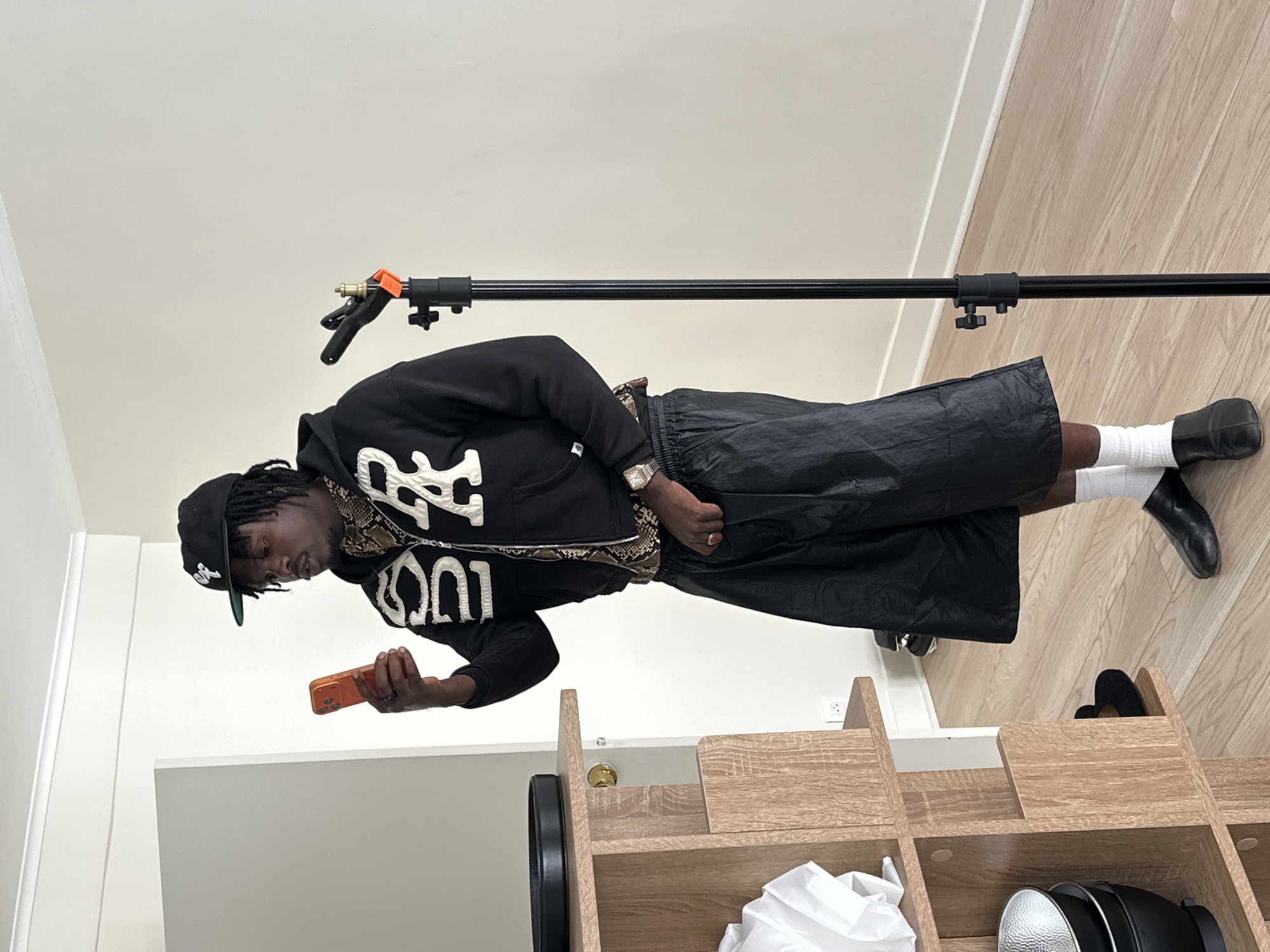 A man in a black hoodie, black cap, and shiny black shorts is standing in an indoor location. He is holding an orange cell phone in his left hand and appears to be looking at it. He is wearing white socks and black shoes. Behind him, there is a wooden shelf unit containing various items, including what appears to be a light fixture and crumpled white material. In the background, a black horizontal pole is suspended. The setting resembles a modern interior, with light-colored walls and wooden floors. The location is not recognizable as being in Paris, France.