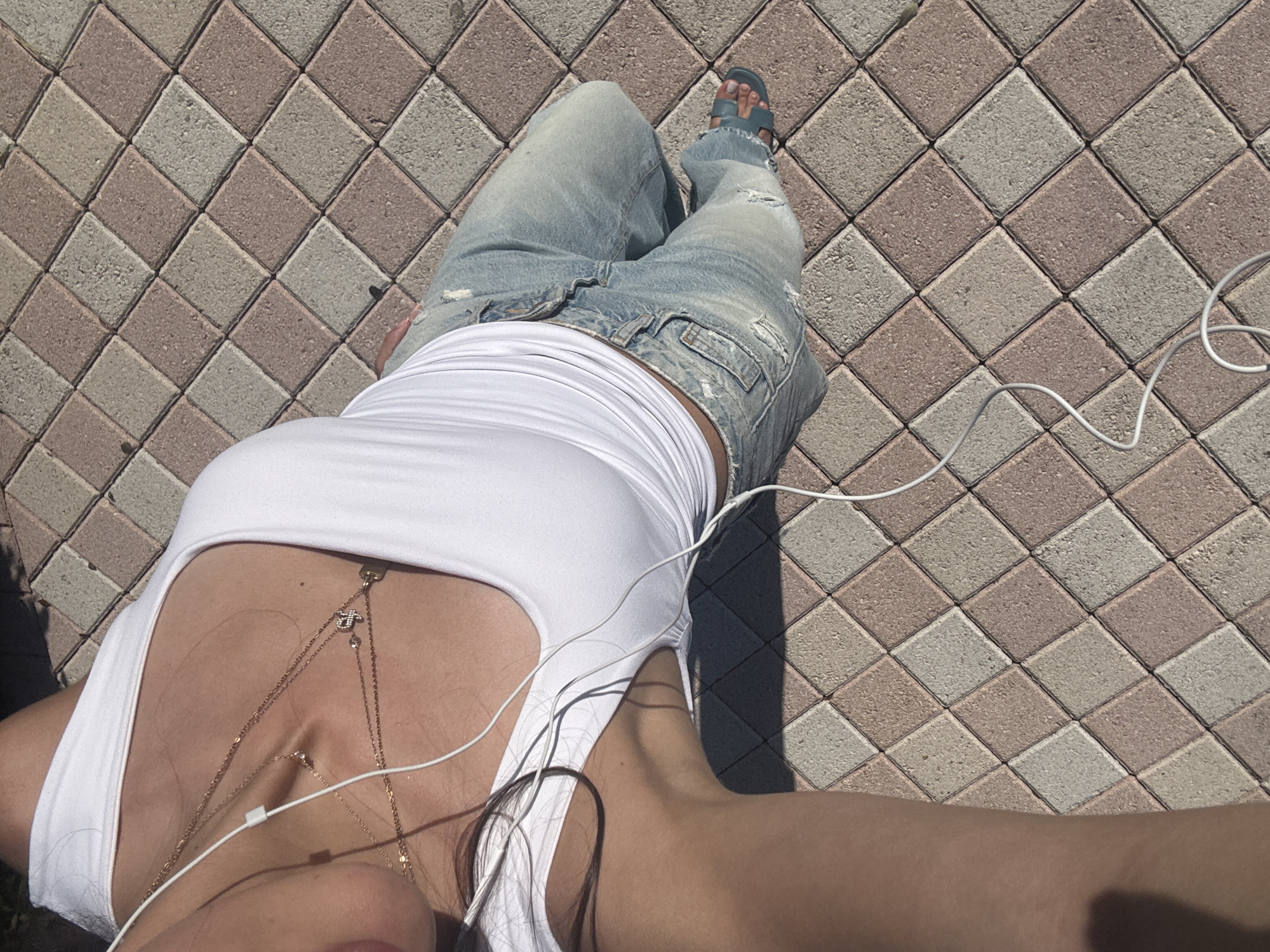 A photograph shows a person from an overhead perspective, lying on a paved surface composed of square blocks arranged in a diamond pattern with alternating colors of light gray and muted reddish-brown. The person is wearing distressed light blue jeans and a white tank top with layered gold necklaces. A white earphone cord is draped across the person's body and the paving stones. A blue sandal is visible on the person's right foot. The location is potentially Boca Raton, United States of America. No specific landmarks or identifying characteristics confirm the exact location.