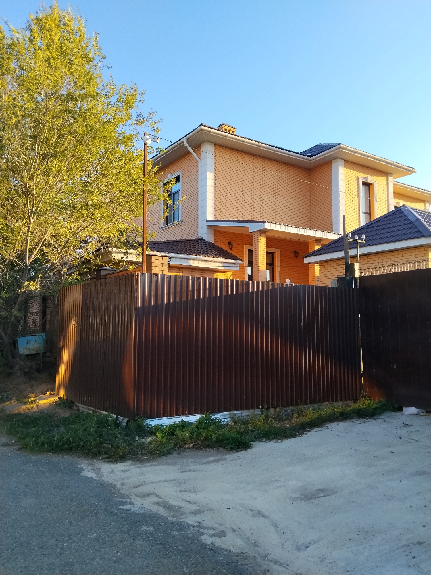 The image captures a scene in Kokshetau, Kazakhstan, featuring a residential property. A two-story house with beige brickwork and white trim is visible behind a tall, dark brown corrugated metal fence. The house has a dark shingled roof and several windows. To the left of the house, a large, leafy tree with bright green foliage stands. A utility pole with a white fixture is positioned near the tree and fence. The ground in front of the fence is a mix of asphalt and concrete, with patches of grass and weeds growing along the fence line. The sky is clear and blue, suggesting a daytime setting.