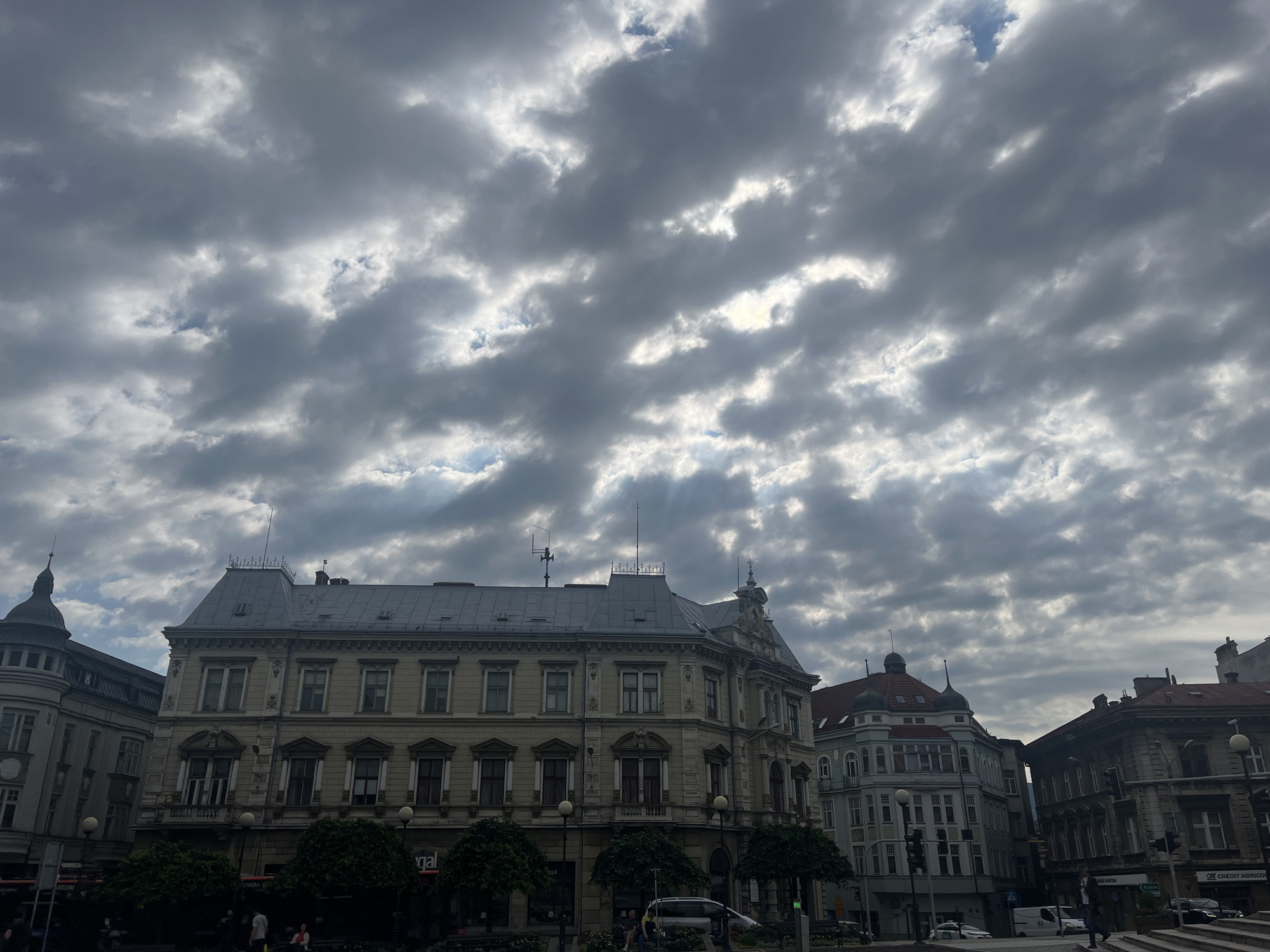 The image captures a cloudy sky over a street scene in a European city. The sky is filled with dramatic, dark grey clouds that are partially illuminated by sunlight breaking through. In the foreground, several ornate buildings with multiple stories line the street. These buildings feature classical architectural details, with numerous windows, balconies, and decorative elements. Trees are planted along the sidewalk, providing some greenery. A few people are visible on the street, some walking and others sitting. Cars and traffic lights are also present, indicating an active urban environment. The overall impression is of a city square or main street on a day with dynamic weather conditions.