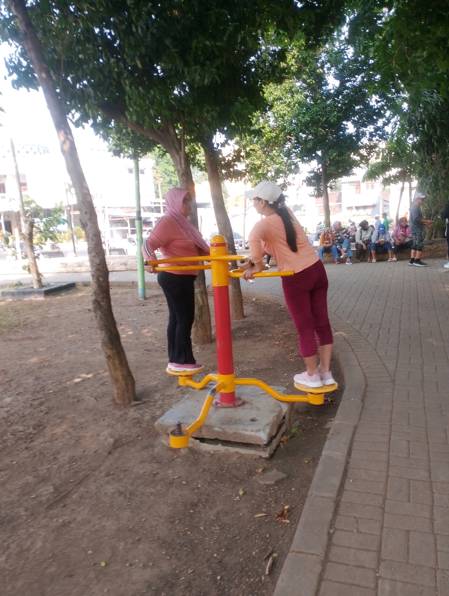 Two women are exercising on an outdoor fitness machine in Probolinggo, Indonesia. The woman on the left, wearing a pink hijab and a pink long-sleeved shirt with black pants and pink sneakers, is standing on the machine's footplates and holding the handles. The woman on the right, wearing a white cap, an orange long-sleeved shirt, and burgundy leggings with white sneakers, is also standing on the footplates and holding the handles. The fitness machine is yellow and red and is bolted to a concrete base. The ground around the machine is covered in dirt and leaves, with several trees providing shade. To the right of the fitness machine, a paved walkway made of interlocking bricks leads away from the scene. In the background, several other people are seated on a bench, and buildings are visible.