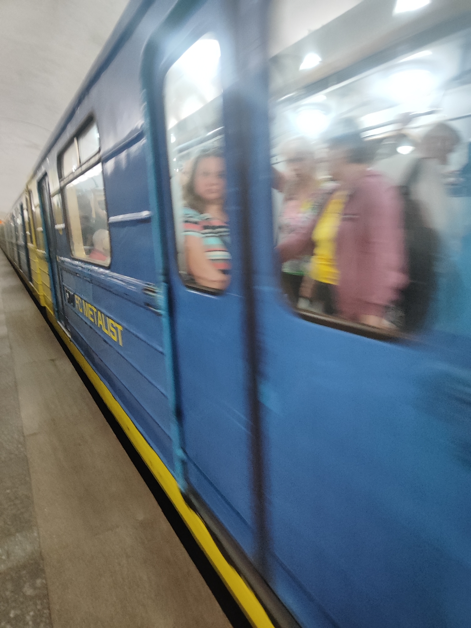 The image shows a blurry view of the interior of a blue and yellow subway train. The train appears to be in motion, as evidenced by the motion blur. Inside the train, several people are visible through the windows. One person in the foreground wears a striped shirt. Other passengers are seen in the background, with some appearing to be talking to each other. The setting is a subway station, with a light-colored ceiling and platform visible. The lighting suggests it is daytime, but no specific time of day can be determined. The overall mood is indistinct due to the blurriness. On the side of the train, the text "FC METALIST" is visible in yellow letters.