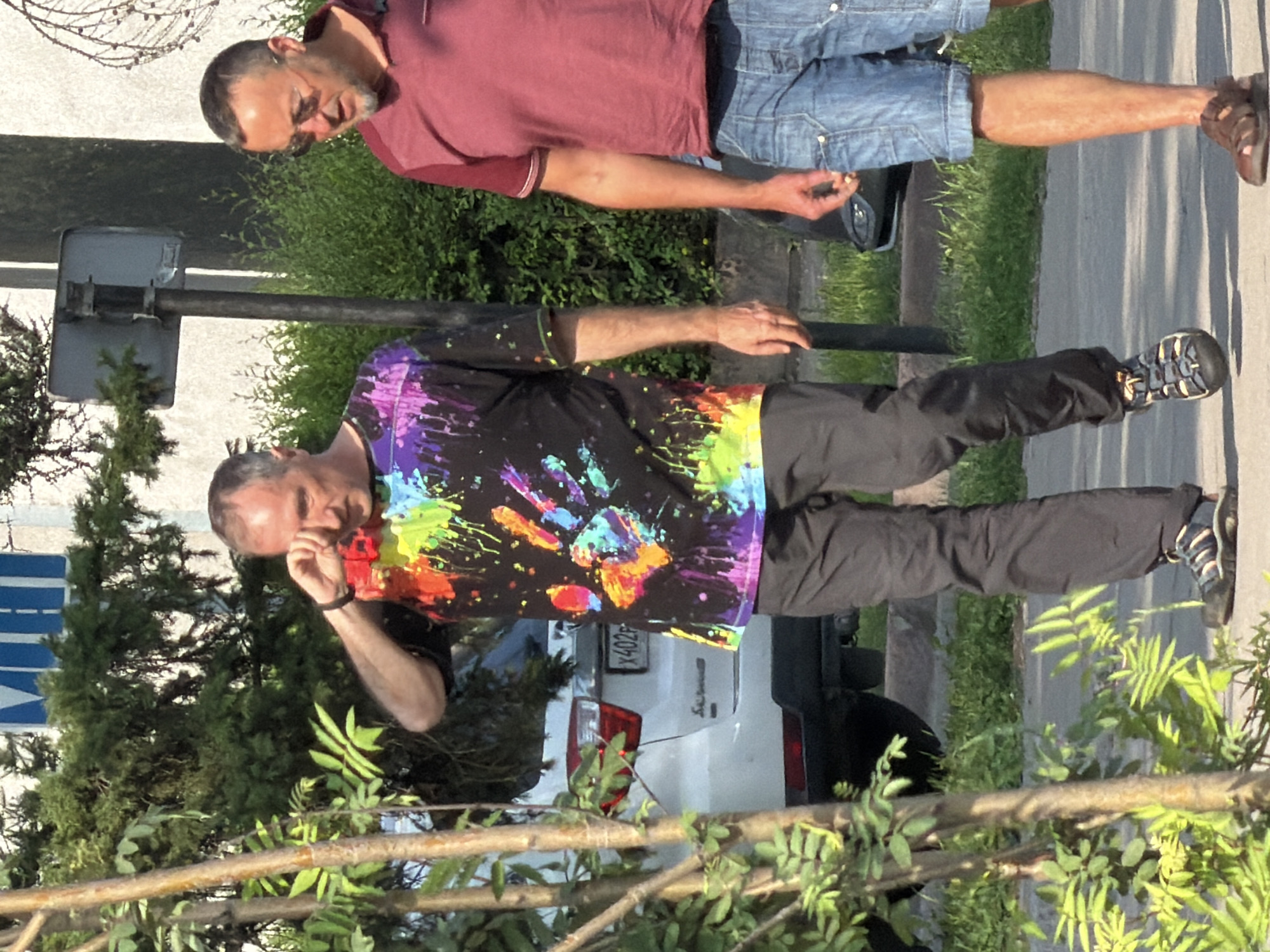 Two adult males are standing on a sidewalk outdoors. The male on the left is wearing a black t-shirt with colorful handprints and splatters, black pants, and dark sandals. He is touching his right eye with his right hand. The male on the right is wearing a maroon t-shirt and denim shorts, and brown sandals. He is holding a cigarette in his right hand. Behind them, a white car is parked. The background consists of green trees and bushes, and a building. The scene appears to be in a city.
