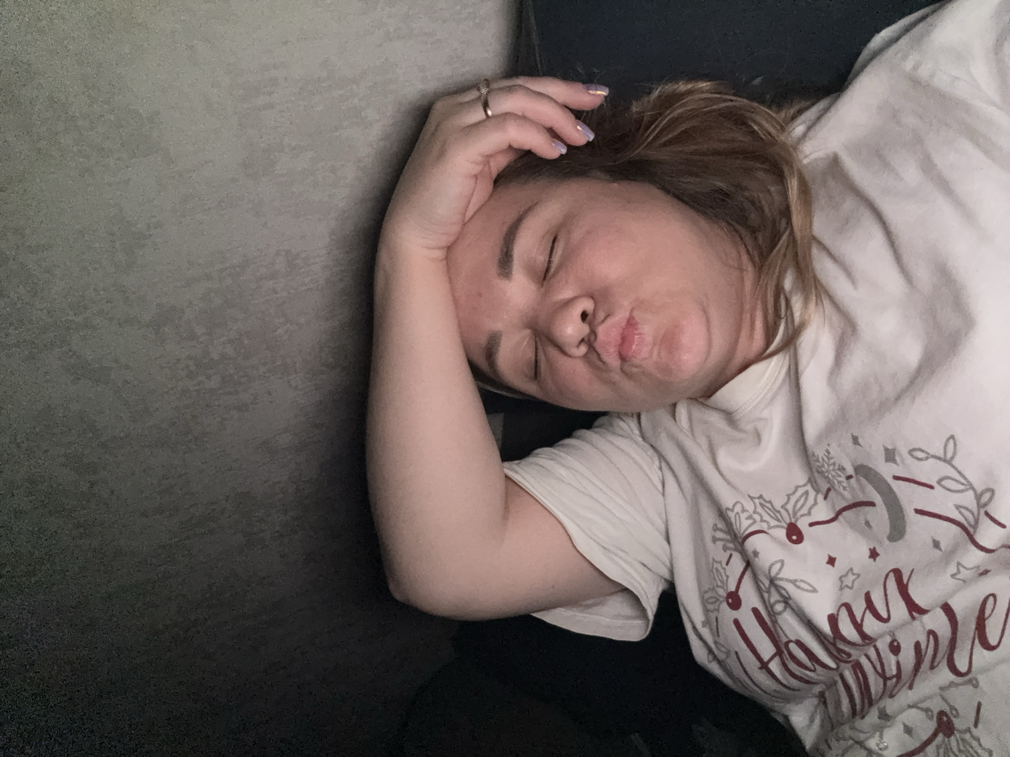 The image shows a woman with her eyes closed, puckering her lips, and resting her forehead on her arm. She is wearing a light-colored t-shirt with a red and grey festive design. A ring is visible on her right hand, which is on her forehead. The background is dark and appears to be a textured wall or surface. The lighting is dim, suggesting an indoor setting at night or in low light conditions. There is no information in the image to suggest the location is in Stary Oskol, Russia.