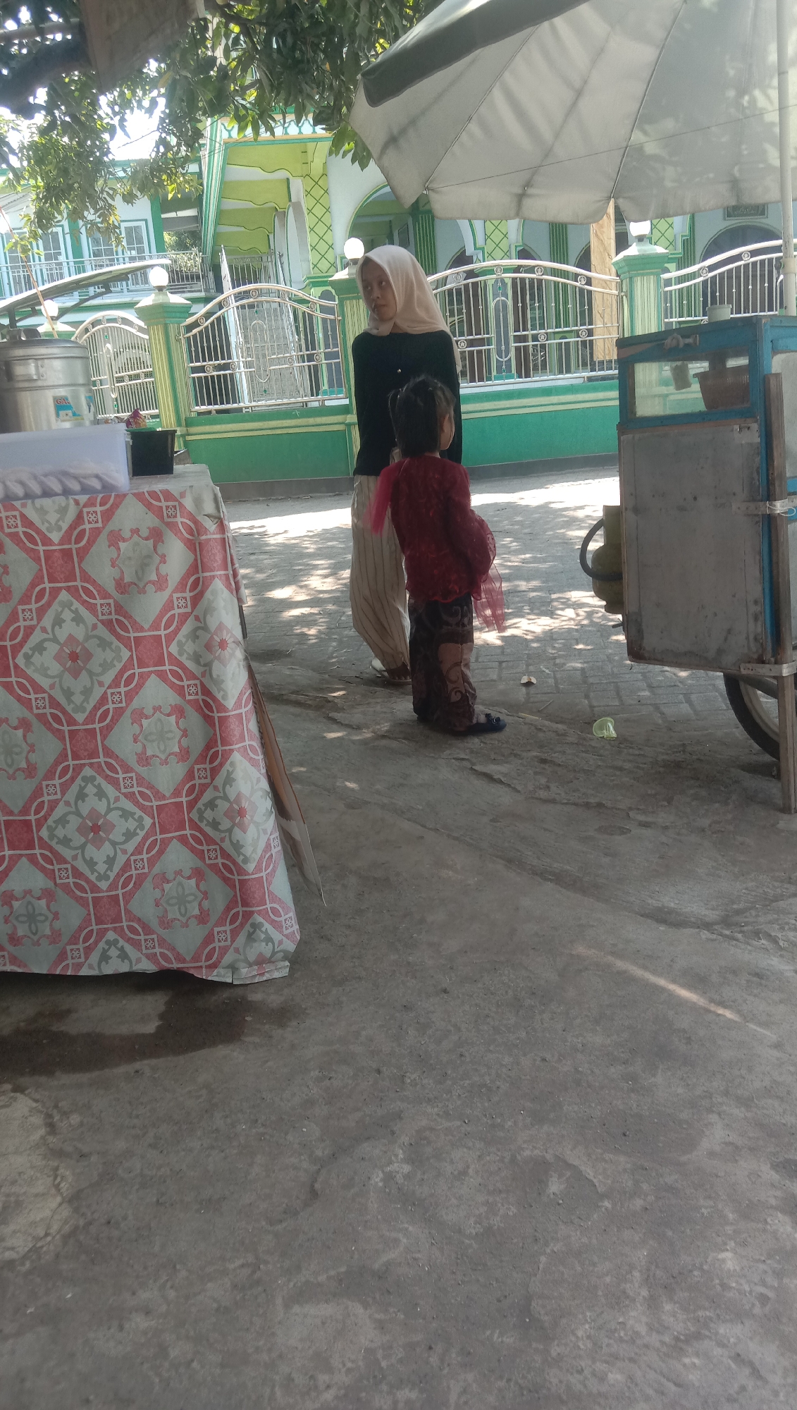 A woman wearing a light-colored hijab and a dark long-sleeved shirt stands beside a young child dressed in a red top and a patterned skirt. They are outdoors on a concrete surface, with a large, patterned tablecloth draped over a table to their left. In the background, a green and white building with arched windows and a decorative fence is visible. A street vendor's cart with a blue frame and metal siding is situated to the right, partially obscured by an umbrella. The scene appears to be in Ngempit, Indonesia, as suggested by the context.
