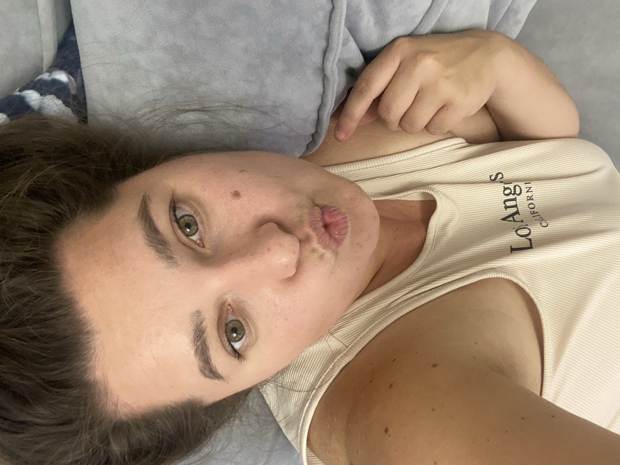 The image shows a selfie of a woman with her mouth puckered, taken from a high angle, likely in a bedroom. She has brown hair pulled back, light green eyes, and fair skin with freckles and a mole on her cheek. She is wearing a cream-colored tank top with "Lo Ang's CALIFORNIA" printed on it. She is lying on a gray surface, possibly a bed or couch. A gray blanket with a blue and white patterned trim is visible behind her head. The lighting appears to be natural, coming from above and to the right. There is no discernible information in the image to suggest the location is the city of Perm, Russia.