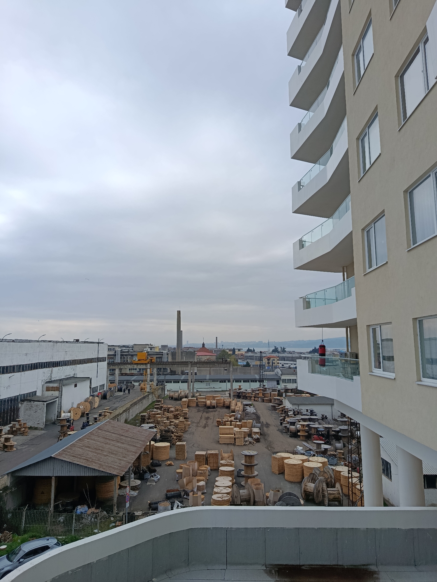 The image depicts an industrial area in Burgas, Bulgaria, seen from an elevated perspective. In the foreground, a modern apartment building with curving balconies stands to the right, its beige facade contrasting with the overcast sky. Below, a large open-air yard is filled with numerous wooden cable spools and packaging. Adjacent to the spools, a shed with a corrugated roof covers stacked cable reels. In the background, industrial buildings line the horizon, punctuated by a tall chimney. Further in the distance, glimpses of the city extend to a body of water. The sky is uniformly gray, contributing to the muted color palette of the scene. A gray car is visible in the lower-left corner.