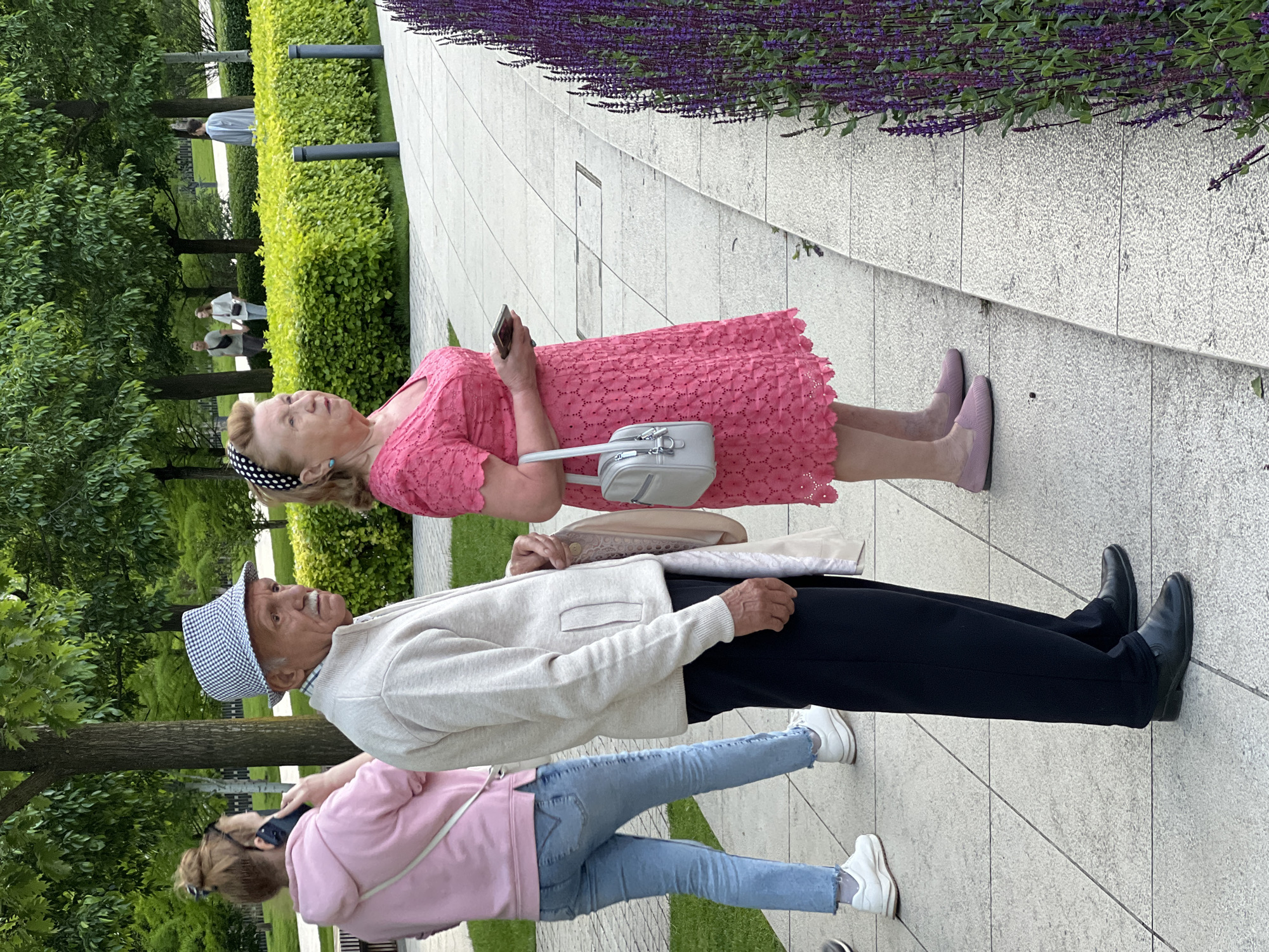 In an outdoor urban setting, three individuals are positioned on a paved walkway adjacent to a grassy area with trees and hedges. An elderly woman in a pink dress, lavender flats, and a polka-dot headband is holding a cell phone and has a light gray purse hanging from her shoulder. Standing beside her is an elderly man wearing a white sweater, dark slacks, black shoes, and a checkered hat. A third individual, appearing younger, is wearing a pink sweatshirt, light blue jeans, and white sneakers, and is also holding a cell phone. The environment includes neatly trimmed hedges, trees, and a row of purple flowers along a wall. In the background, several individuals are walking, suggesting a public park or garden. Based on the description and image content, this location is most likely in the city of Krasnodar, Russia.