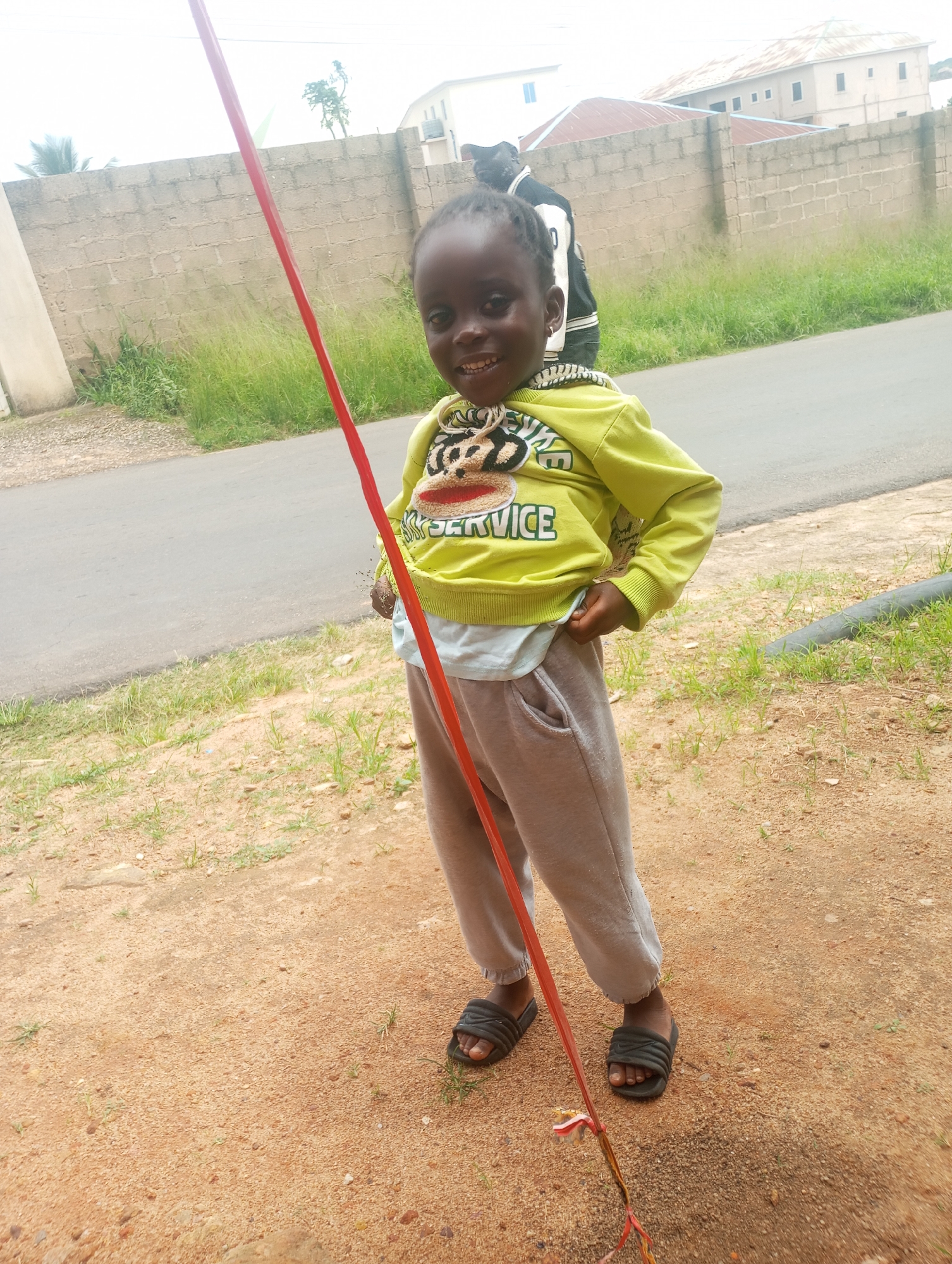 A young Black child with dark skin, braided hair, and a bright smile stands outdoors. The child is wearing a lime green sweatshirt with a graphic print and light gray sweatpants, along with black slip-on sandals. A long, thin red object is held by the child and extends upwards, partially obscuring the view. In the background, a beige brick wall with green grass and a road is visible. Behind the wall, there are buildings with red roofs. The ground in front of the child is composed of reddish-brown sand and scattered pebbles with sparse green grass. The overall impression is a candid outdoor shot in a residential area.