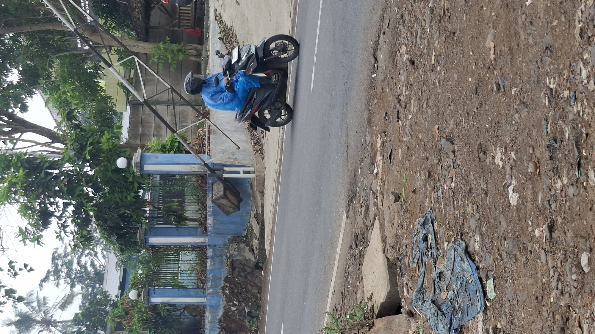 In Cilaku, Indonesia, a person wearing a blue rain suit and helmet rides a black motorcycle on an asphalt road. To the left of the road, a blue and white striped fence encloses an area with trees and a concrete wall. A pile of dark rocks and debris sits near the fence. A metal structure, possibly part of a gate or support, extends diagonally across the upper right portion of the image, with some signage visible in the background. The ground in the foreground is covered with rocks, dirt, and scattered blue tarp and other debris.