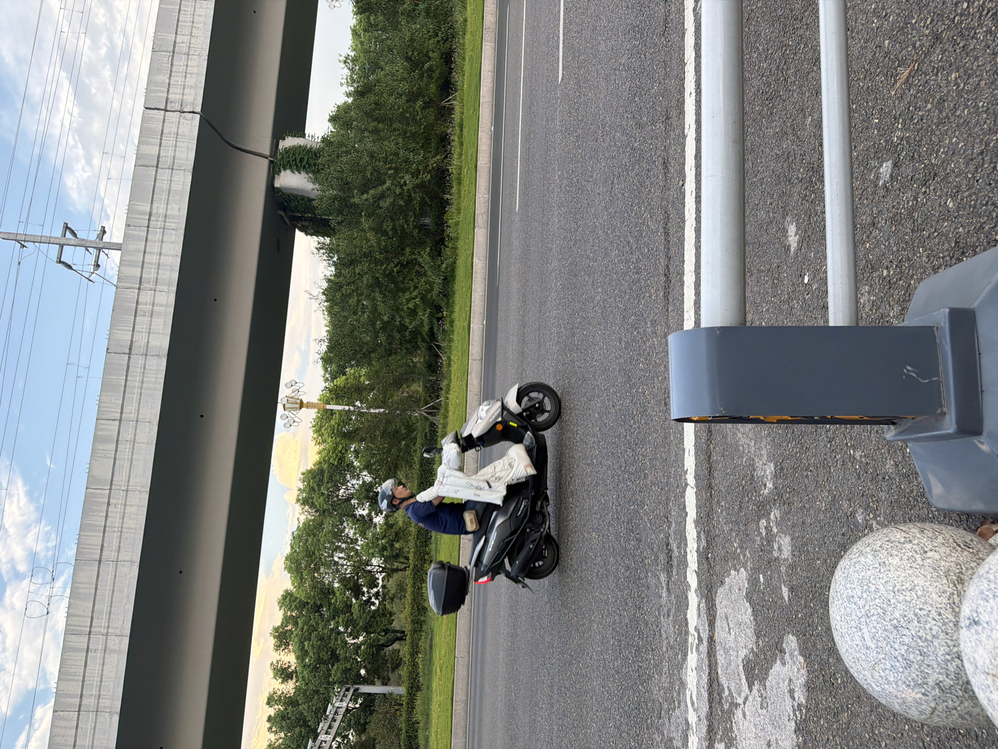 A person rides a scooter on a road in Jiangjin District, China. The person is wearing a blue shirt, dark pants, and a helmet. They are carrying a white object in front of them on the scooter. The scooter has a black storage box on the back. In the background, there is a large elevated concrete structure with visible cracks, supported by pillars. Trees and greenery are visible behind the road. The sky is partly cloudy with some yellow hues, suggesting it may be late afternoon or early evening. A metal guardrail is in the foreground.