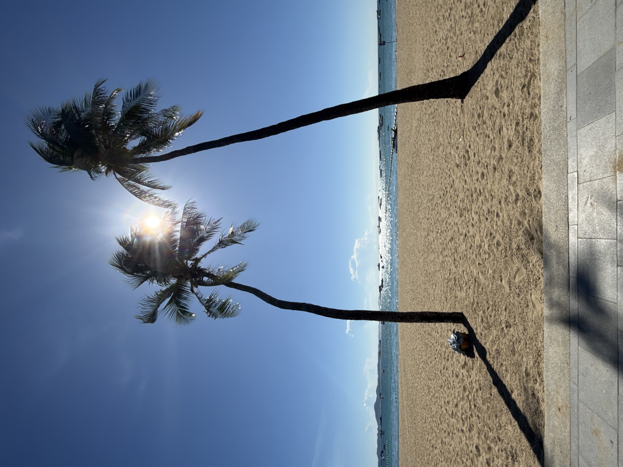 The image depicts a bright, sunny day on a sandy beach in Pattaya City, Thailand. Two palm trees stand prominently in the foreground, silhouetted against a clear blue sky and the radiant sun, which is partially obscured by the fronds of the tree on the left. The sun's rays create a lens flare effect. The sand is smooth with visible footprints. In the background, the calm turquoise ocean meets the horizon, dotted with several boats and a marker buoy line. The shore is lined with a paved walkway in the very bottom foreground. A small bundle of items, possibly luggage, rests at the base of the left palm tree, casting a long shadow. The overall scene is serene and evokes a typical tropical beach atmosphere.