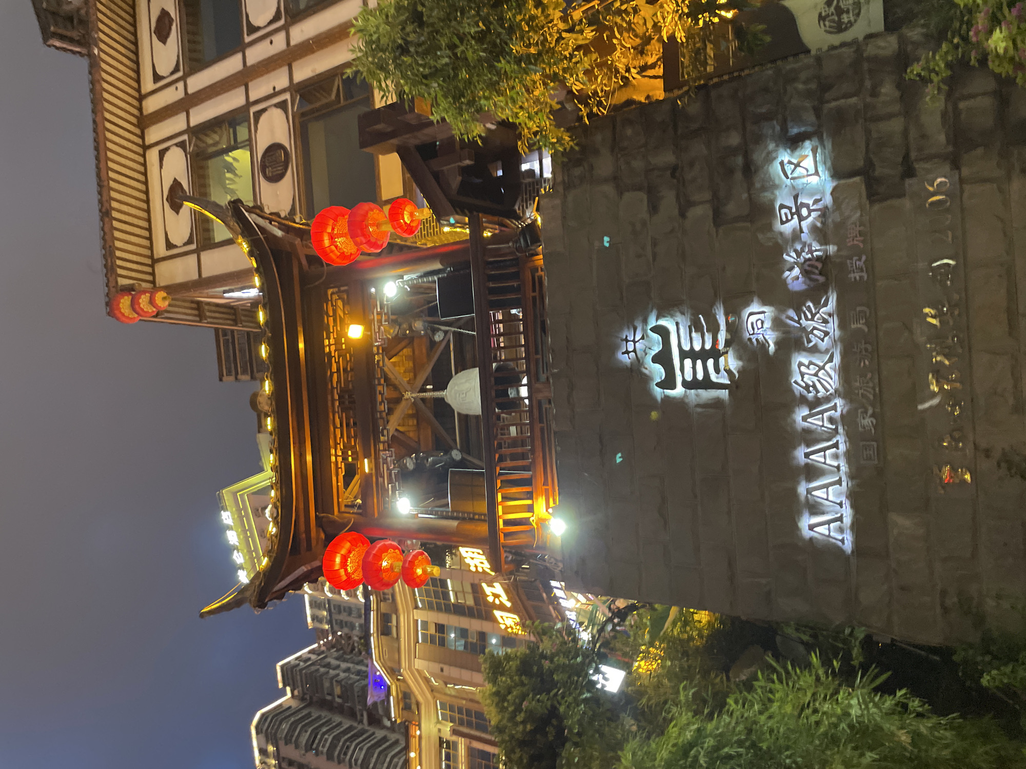 The image shows an outdoor nighttime scene in Yuzhong District, China, featuring a traditional Chinese pavilion adorned with red lanterns. The pavilion has a tiered roof with intricate detailing and is illuminated by warm lighting. Below the pavilion, a stone wall displays illuminated Chinese characters and the text "AAAAA 级旅游景区" (AAAAA Level Tourist Attraction) in English. The background shows modern buildings with illuminated windows, suggesting an urban environment. Green foliage frames the scene on the left and right.