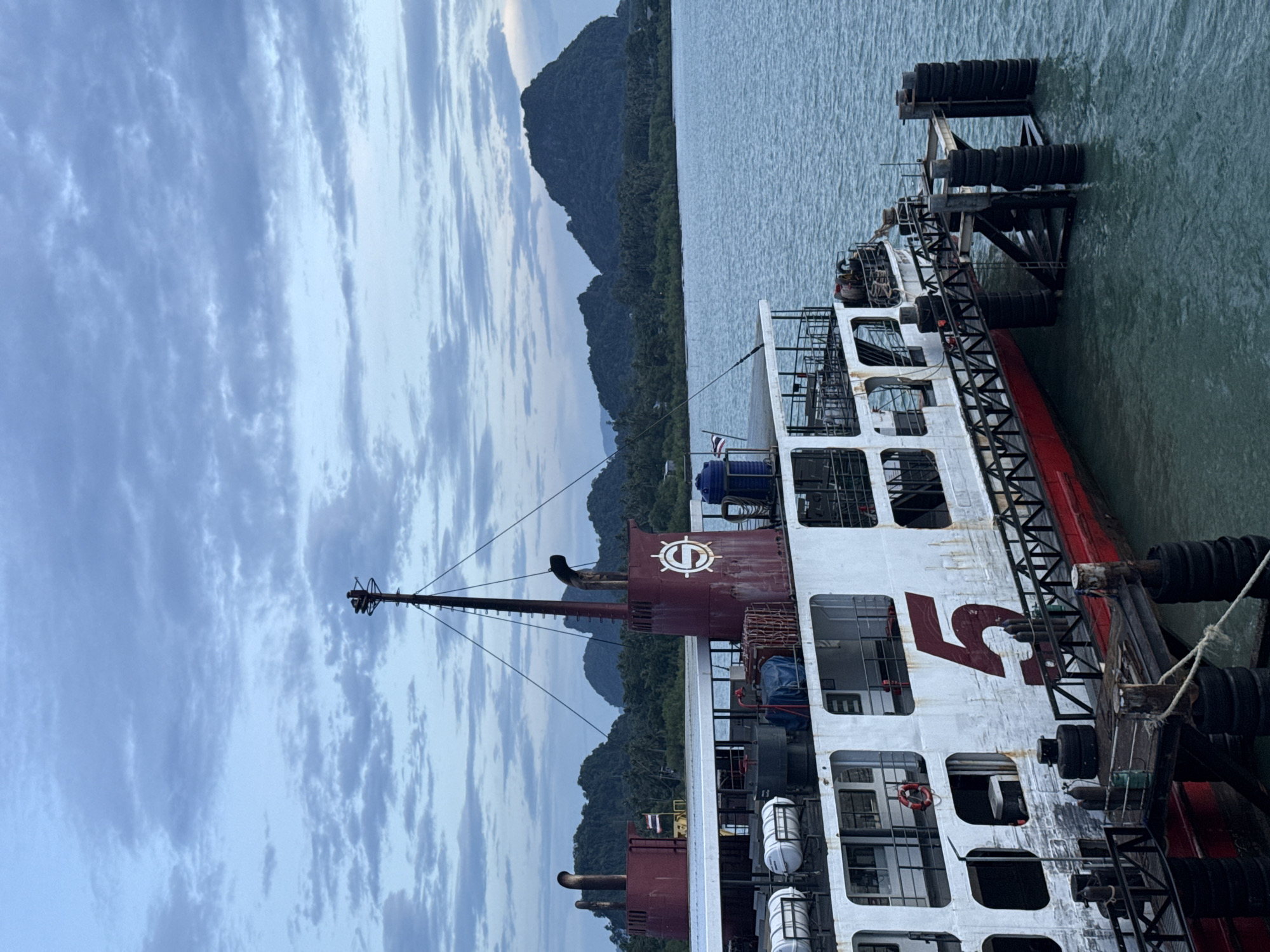 This image shows a ferry docked at a pier in Thailand. The ferry is white with a red hull and a large red number "5" on its side. It has several decks and lifeboats are visible on the upper deck. The ferry is connected to a pier with a gangway. The pier is supported by pilings wrapped in tires. In the background, there are lush green islands with palm trees. The sky is cloudy and a bit overcast. The water is a calm, greenish-blue. There are no people clearly visible in the image. The scene appears to be at dusk or dawn. The location is identified as a ferry terminal in Thailand, though a specific city is not mentioned.
