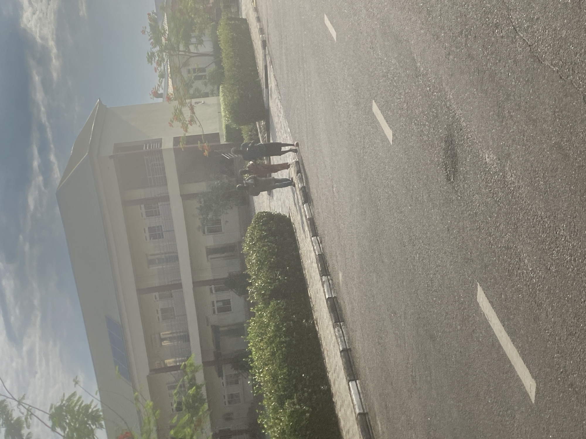 A daytime shot in Angwan Sayawa, Nigeria, shows a building with multiple floors and windows on the left. In front of the building is a line of green bushes along the sidewalk. On the right is an asphalt road with white lines painted on it. Several individuals are walking on the sidewalk. The sky is visible in the upper left corner with clouds. The image appears to be taken from a slightly elevated position.