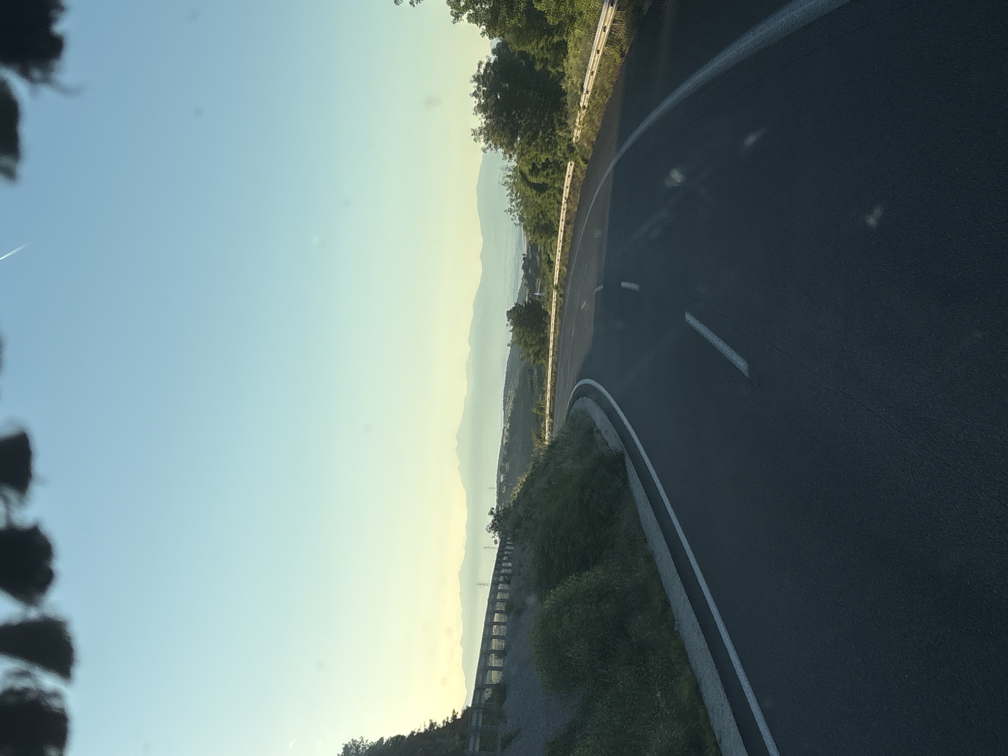 The image depicts a winding asphalt road curving to the right, bordered by a metallic guardrail and lush green vegetation on the left. In the distance, a range of hazy mountains stretches across the horizon under a clear, pale blue sky. The scene appears to be captured from a moving vehicle, as indicated by the blurred dark shapes at the top of the frame, suggestive of a windshield or roofline, and a faint contrail visible in the sky. The lighting suggests either early morning or late afternoon, with a soft, diffused sunlight. The location is identified as Avra, Greece.