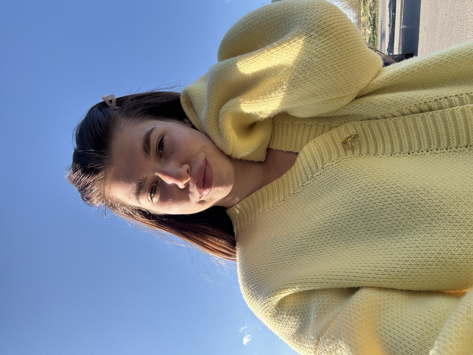 A young woman is pictured from a low angle against a bright blue sky. She is wearing a pastel yellow knit sweater with a star-shaped pin on the right side of the collar. The sweater appears to be bulky, with a portion draped over her shoulder. Her brown hair is styled with a clear hair clip on the left side of her head, pulling back some of her hair. Her face is slightly turned towards the camera, with a soft expression. The background is primarily sky, with a hint of buildings and trees visible in the upper right corner, possibly in the city of Речицы, Russia.