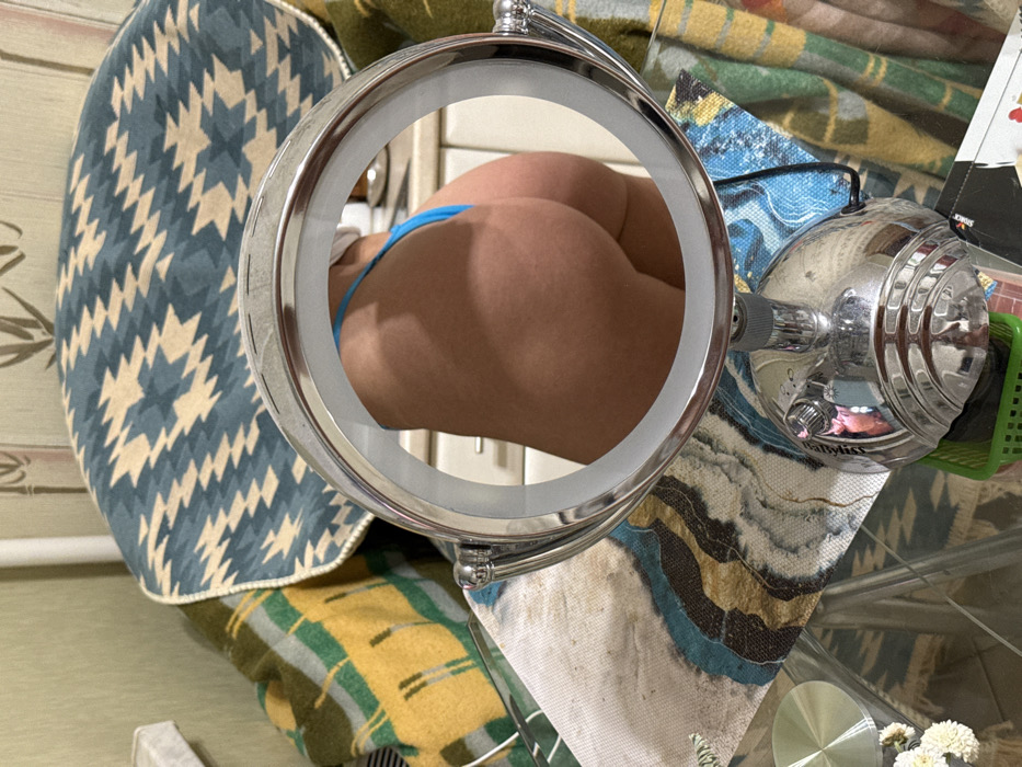 An individual's buttocks, partially framed by a round mirror, are the central focus. A thin blue thong is visible. Surrounding the mirror is a mix of textures and patterns: a pillow with a geometric design, a patterned blanket, and a cluttered surface with a hair styling device. The scene is indoors, illuminated with a bright, even light.