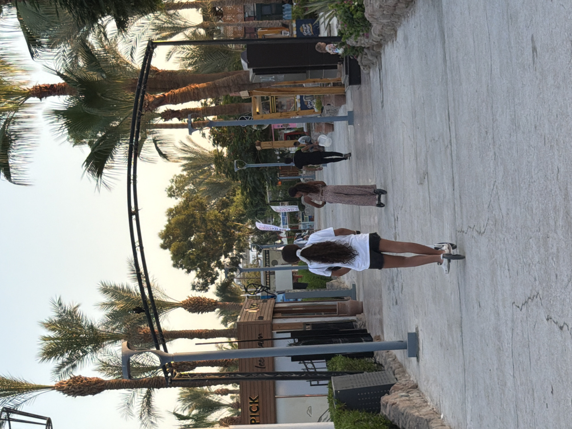 In a pedestrian walkway in Sharm El Sheikh, Egypt, a young girl with long, dark, curly hair tied in a ponytail rides a white scooter away from the camera. She is wearing a white t-shirt and dark shorts. To her right, a woman in a patterned jumpsuit also rides a scooter, moving in the same direction. Further ahead, a man in a black t-shirt and pants walks away from the camera carrying a white bag. Several palm trees line the pathway, with a metal archway extending overhead. Buildings with wooden facades and signs are visible on the left side of the walkway. The scene appears to be set in the late afternoon or early evening, with soft lighting.