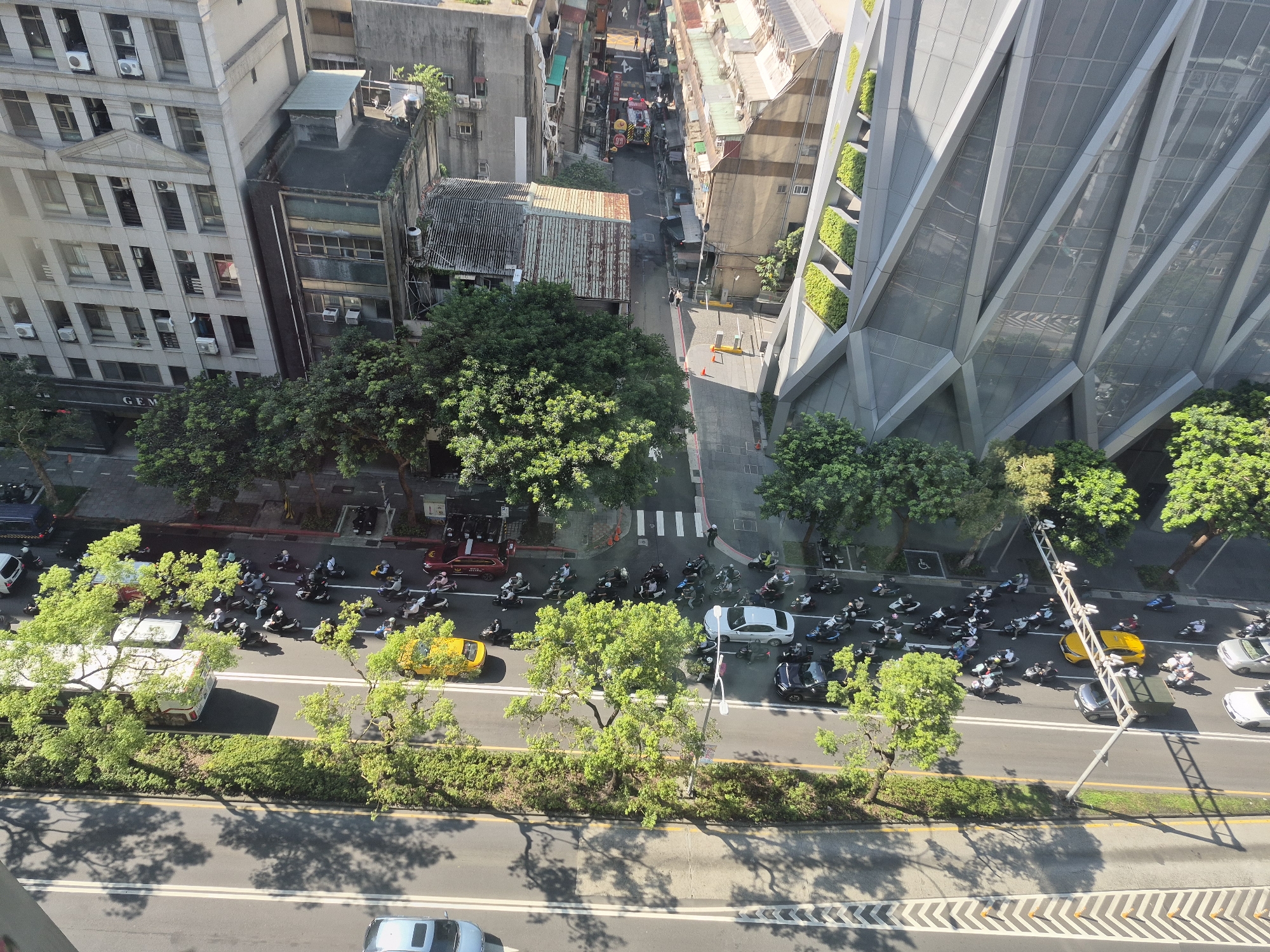 The image provides an overhead view of a busy street scene in Taipei, Taiwan. On the left side of the frame, a tall, light-colored building with many windows and air conditioning units is visible. Adjacent to it are smaller, older buildings with corrugated metal roofs. A dense canopy of green trees lines the street, casting shadows on the asphalt.
The street itself is filled with a large number of motorcycles and scooters, with a few cars and a white van. The vehicles are in motion, suggesting traffic. A bright yellow car is particularly noticeable. The road features white lane markings and a pedestrian crossing.
In the background, a modern building with a striking geometric facade and integrated green spaces stands prominently on the right side of the image. The architecture is sleek and contemporary, contrasting with the older buildings. Further back, more urban structures and smaller streets can be seen. The overall impression is of a dynamic urban environment with a mix of old and new architectural styles, and a high volume of vehicle traffic.
