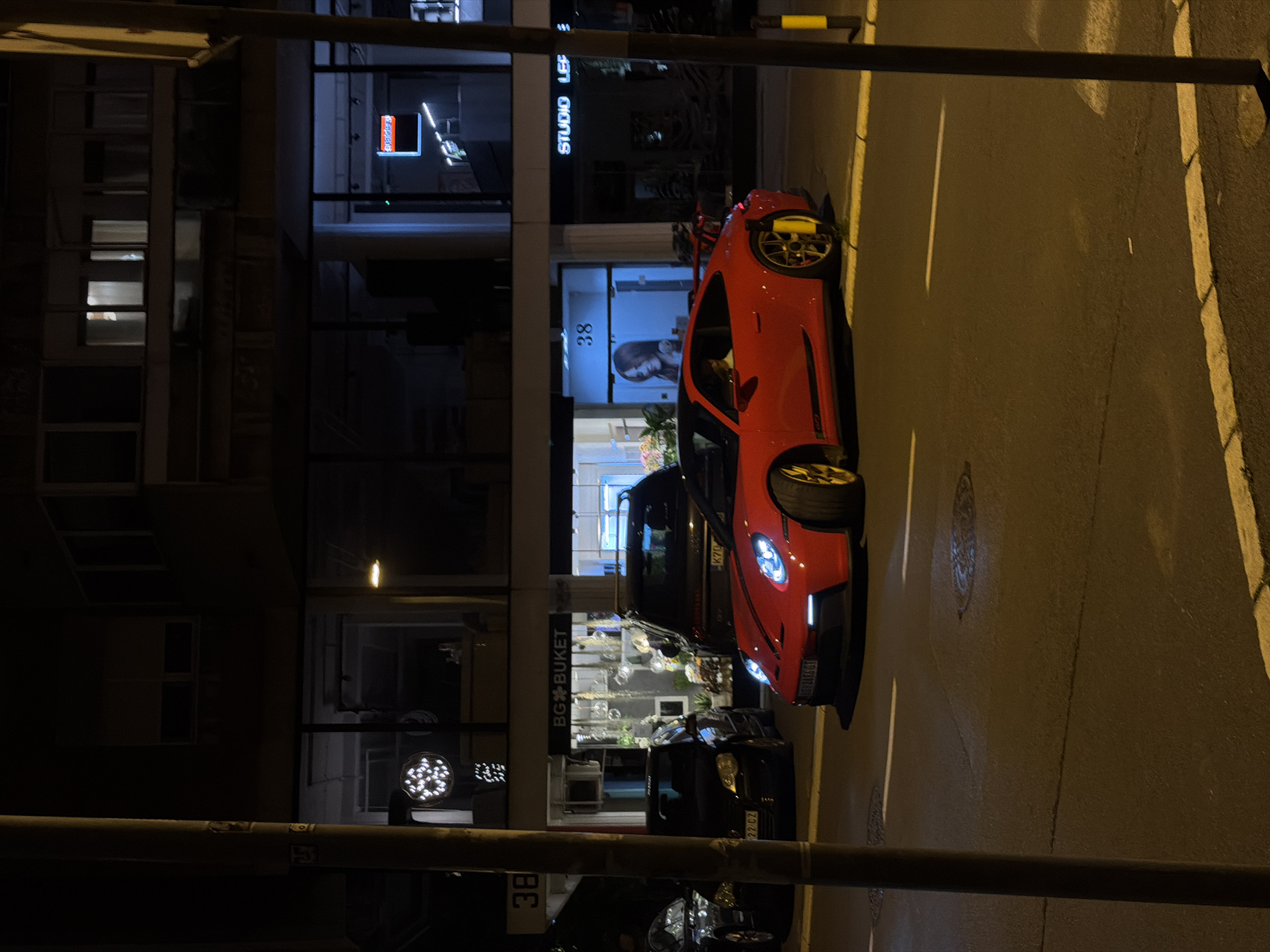 This image captures a nocturnal scene on a city street, likely in Vracar Urban Municipality, Serbia, given the visible Serbian license plates and store signage. In the foreground, a vibrant red Porsche sports car with yellow accents on its wheels is parked on the asphalt road. The car's headlights are illuminated. Behind the Porsche, a dark-colored SUV and another black car are also visible, parked on the street. The background features the facade of a building with multiple windows and storefronts. One storefront displays the name "BG BUKET" with an arrangement of light fixtures and plants inside. Another storefront has a blue sign with the number "38" and an image of a woman. Further along the building, the words "STUDIO LEF" are discernible on a sign. The street is illuminated by streetlights, casting a warm glow on the scene. The overall impression is that of a quiet night in an urban setting, with a focus on the presence of parked vehicles and the illuminated storefronts.
