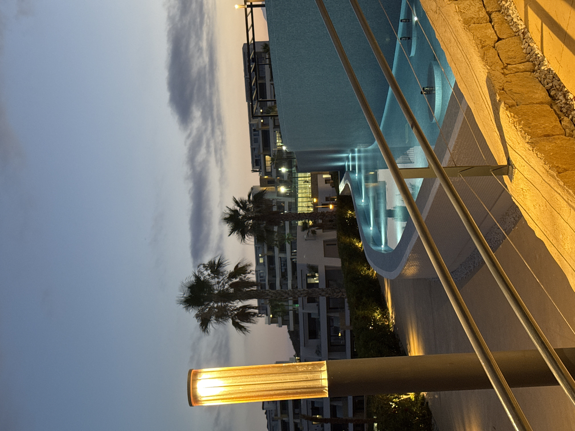The image depicts a twilight scene at a resort in Finestrat, Spain. In the foreground, a tall, illuminated cylindrical lamp with a textured glass exterior stands to the left, casting a warm yellow light. A metal railing with multiple horizontal bars follows a curved path downwards from the right foreground. Beyond the railing, a blue swimming pool is visible, with sections featuring submerged illuminated circular platforms. The pool is surrounded by a light blue tiled wall that curves along its edge. In the mid-ground, palm trees with prominent fronds are interspersed with lush green bushes. Behind the vegetation, modern multi-story buildings with balconies and illuminated windows form the backdrop. The sky above is a gradient of deep blue transitioning to lighter hues towards the horizon, with some scattered clouds. The overall ambiance suggests a peaceful evening at a well-lit resort.