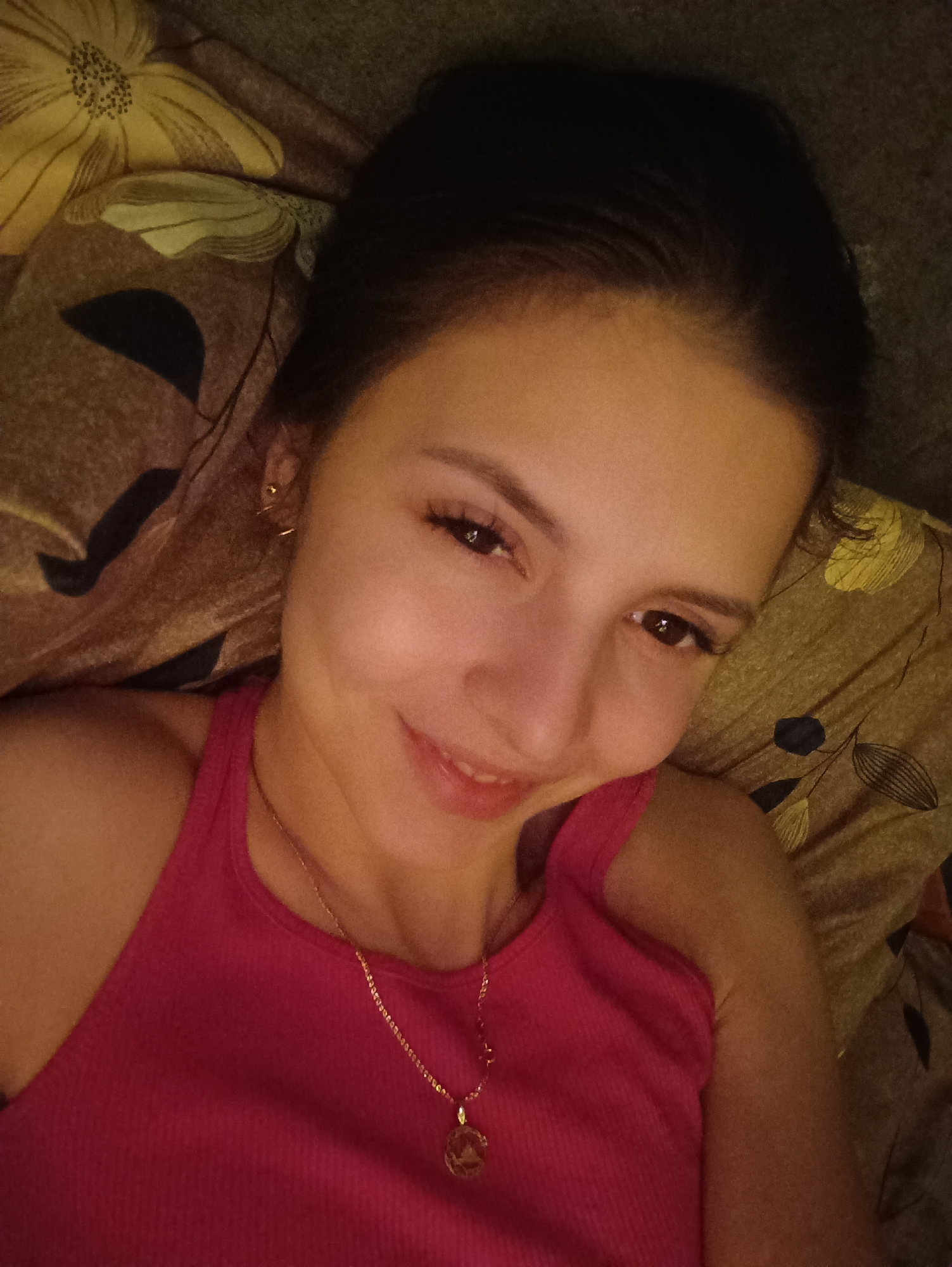 A woman is lying down, looking at the camera with a slight smile. She is wearing a pink tank top and a gold necklace with a round pendant. She has dark hair pulled back and is on patterned bedding, with flowers and leaves in dark hues. The location is Unknown, Unknown.