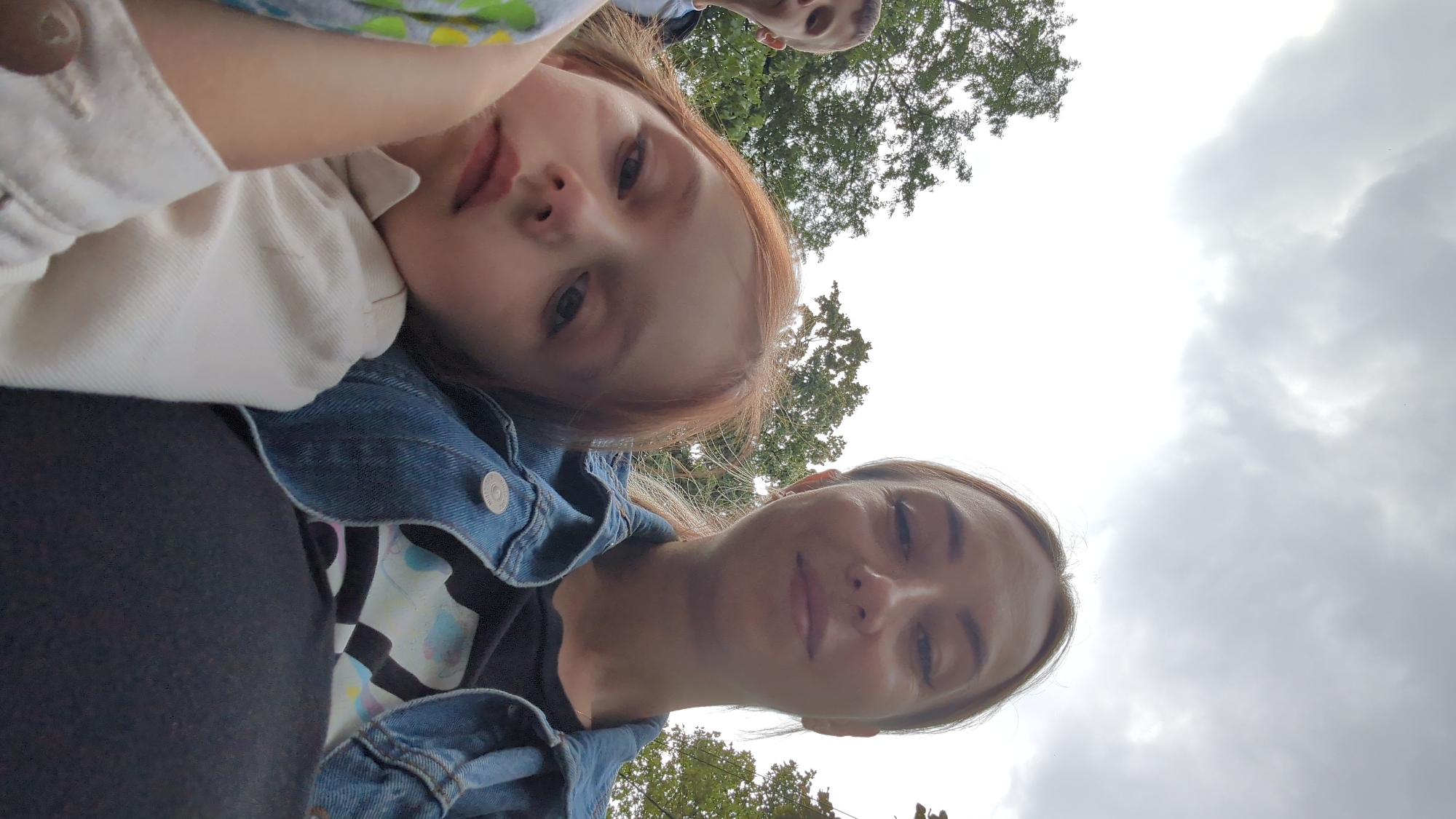 A selfie shows a woman and two children outdoors. The woman is in the foreground on the right, wearing a denim jacket over a black t-shirt. Her hair is pulled back. To the left of the woman, a young girl with red hair rests her head on the woman's arm. She is wearing a white top. Partially visible to the far left is a young boy, also wearing a light-colored top. The background features trees and a cloudy sky. The image was taken in Chișinău, Moldova.