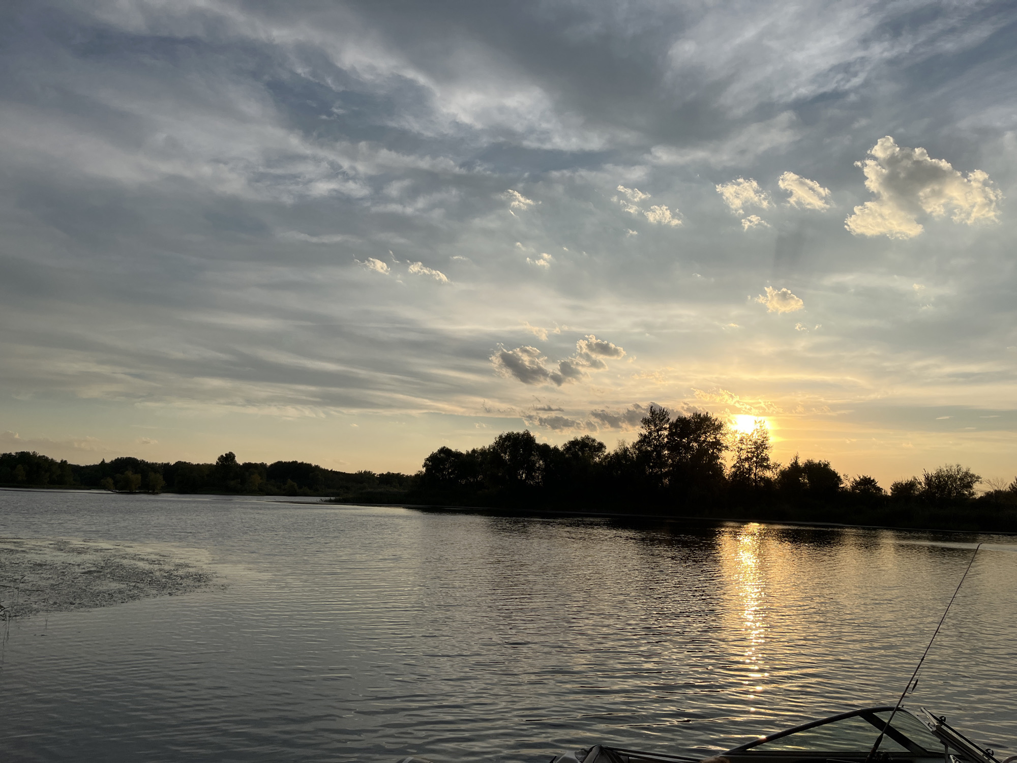 A body of water, likely a river or lake, is depicted with a sunset in the background. The sky is filled with streaks of clouds, with the sun peeking through on the right side, casting a golden reflection on the water. A dark silhouette of trees lines the opposite shore, creating a stark contrast against the illuminated sky and water. In the foreground, the front of a boat is visible, with a fishing rod extending upwards. The water has a rippling texture, indicating a slight breeze. The scene suggests a tranquil evening, possibly a fishing excursion, in a natural setting. The provided location of Шумейка, Russia, places this scene in a specific geographical context.