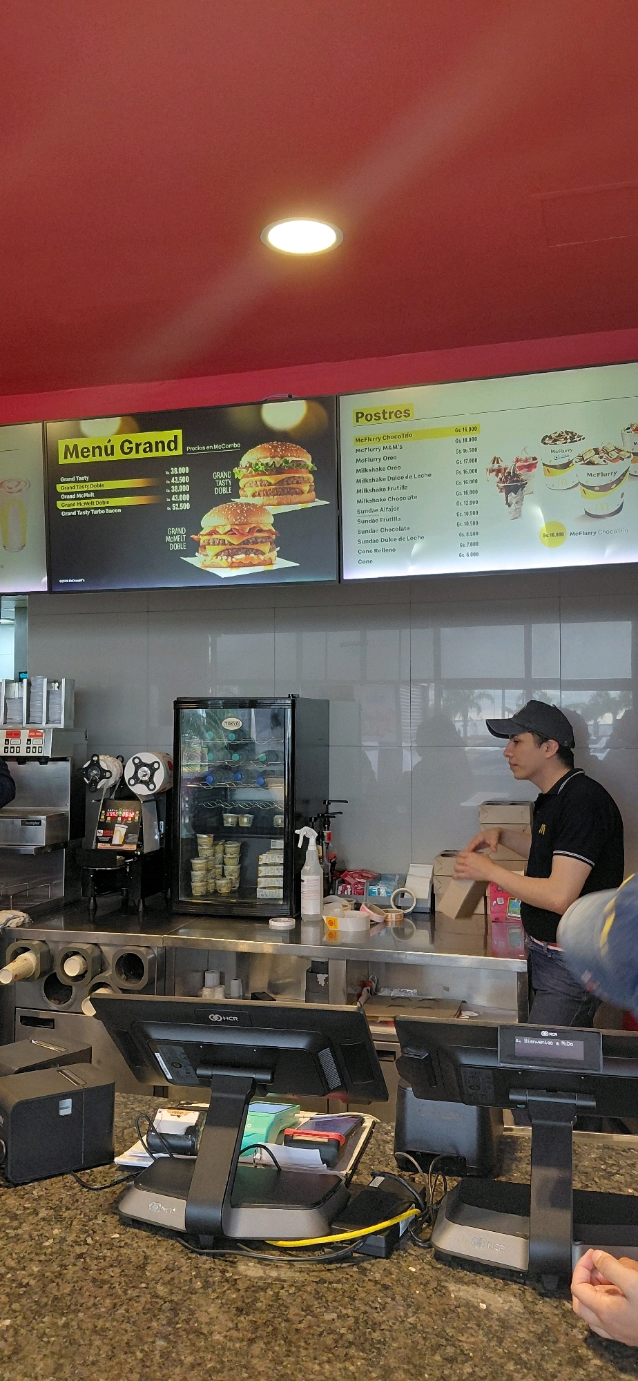 The image captures an interior view of a McDonald's restaurant, likely at the location in Encarnacion, Paraguay, given the menu item names and currency. The scene is dominated by the counter area, with a prominent menu board displaying "Menú Grand" and "Postres" (Desserts) with prices listed in Guaraníes (Gs.). Two large images of burgers are featured on the "Menú Grand" board, showcasing the "Grand Tasty Doble" and "Grand McMeltt Doble."
On the right side of the frame, a male employee wearing a black McDonald's uniform and a dark baseball cap is standing behind the counter. He is looking to his right and appears to be engaged in an activity involving cardboard boxes. Several boxes are stacked on the counter in front of him, and he is holding one of them.
In the background, a glass-door refrigerator unit is visible, stocked with drinks and what appear to be small containers of food. Various operational equipment, such as a drink dispenser, a receipt printer, and touch-screen point-of-sale (POS) systems, are arranged on the counter. The overall environment suggests a busy fast-food establishment during operating hours. The ceiling is red, and a circular light fixture is visible.
