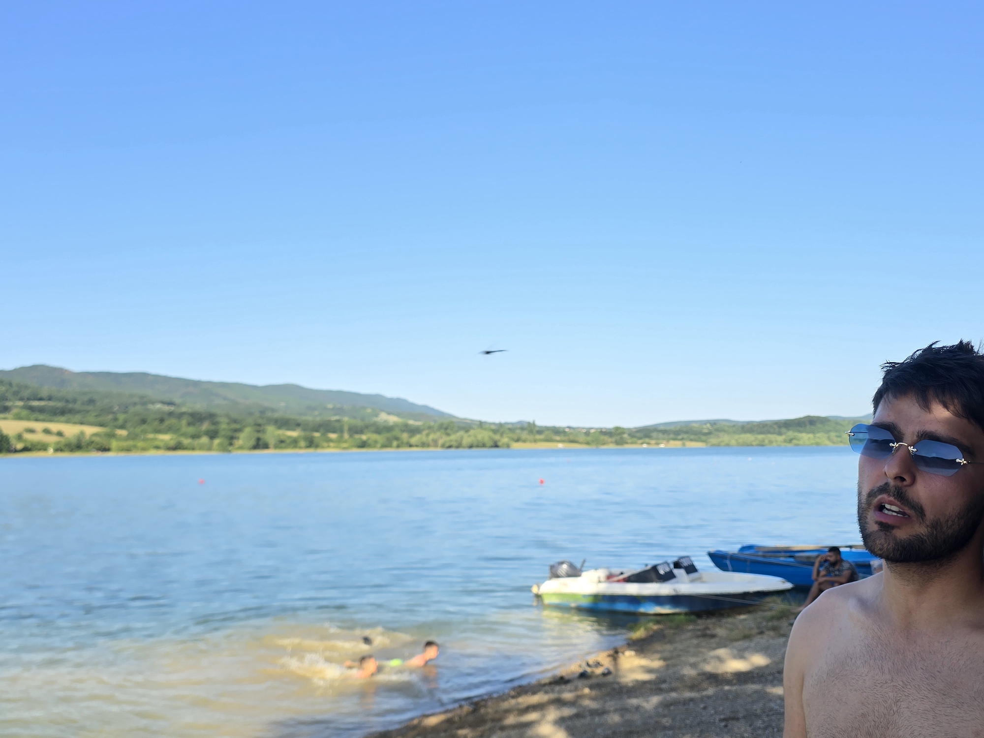 A man wearing blue-tinted sunglasses and no shirt is in the foreground on the right, looking to his left with his mouth slightly open. Behind him, on the shore of a lake, are several boats. Two are blue, and one is white and green with a motor. In the water to the left, two children are swimming, creating splashes. The lake is blue, and across it, a tree-lined hilly landscape is visible under a clear blue sky. The location is identified as the city of Golyama Zhelyazna, Bulgaria.