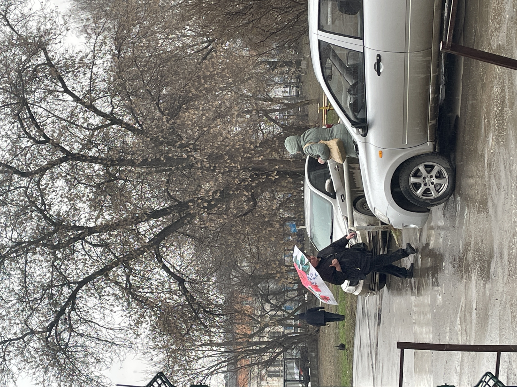 On a rainy day in Izhevsk, Russia, an individual holding a floral umbrella walks past parked cars. A second person is visible at an open car door. Trees with bare branches are in the background.