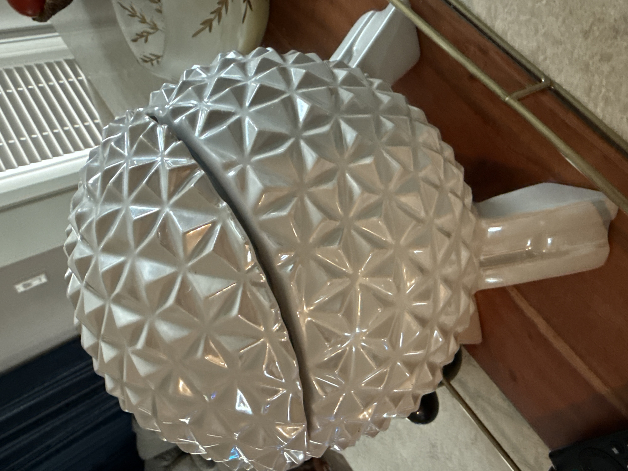 The image shows a silver-colored, decorative object resembling a pineapple sliced in half, sitting on a wooden surface. The pineapple has a textured surface consisting of small, raised triangular shapes. It features stem-like projections on each half. The object is located indoors, near a window with horizontal blinds, and possibly on a table with a metal railing. The location appears to be a residence or commercial building in Orlando, United States.