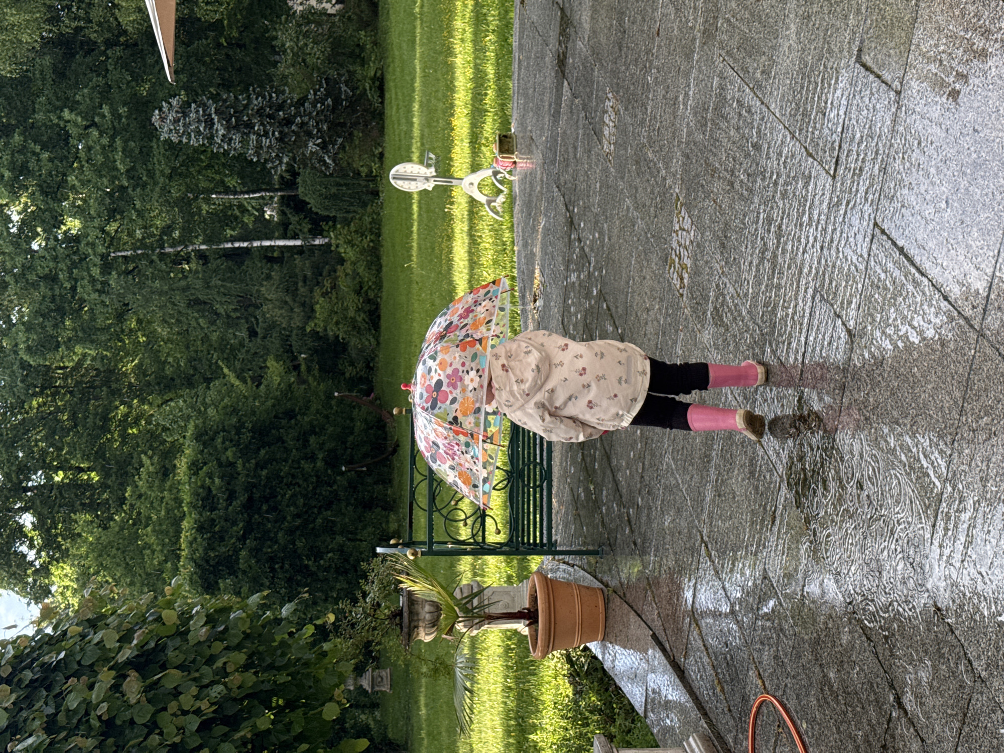 A girl with pink boots and a flowered umbrella stands on a wet, grey stone patio, beside a bench and potted plant. The background shows a green lawn and trees. The scene appears to be in a garden. There is no information to suggest that the location is in the city of Sapronovo, Russia.