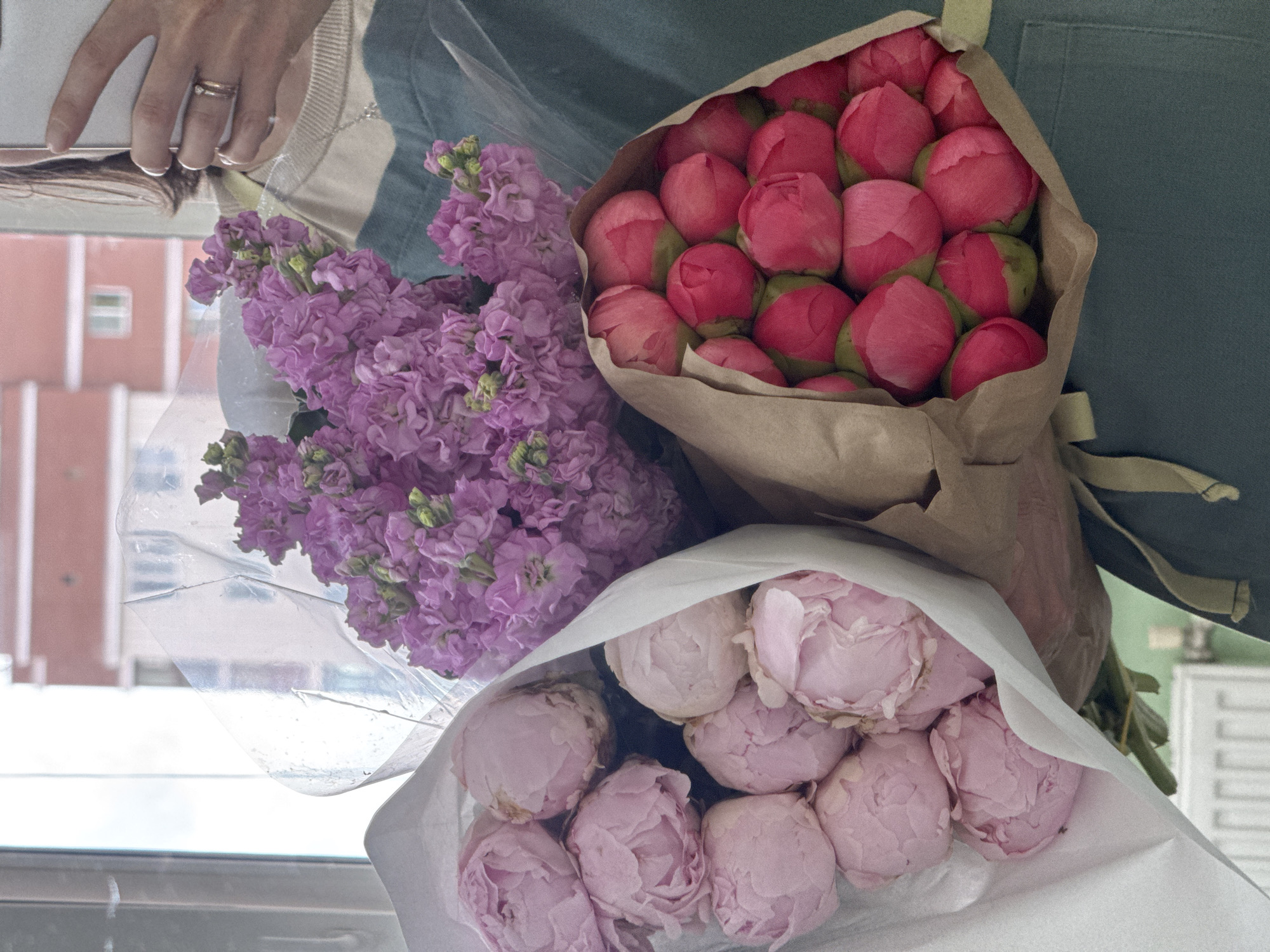 The image shows a hand holding three bouquets of flowers. The first bouquet contains purple flowers with green stems. The second bouquet contains pink peony buds wrapped in brown paper and tied with a light green ribbon. The third bouquet contains pink peonies wrapped in white paper. The hand is wearing a silver ring. The background shows a building with a pink facade and white window frames. Based on the style of the building, it is possible that the location is Izhevsk, Russia. The image is lit by natural light coming from a window.