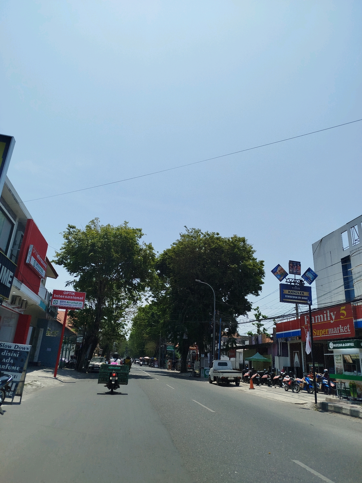 The image shows a street scene in Kebumen, Indonesia, on a bright, clear day. The road is asphalt, with a white line marking the center. On the left side of the road, there is a building with red and white signage, including "Optik Internasional" and a sign that reads "SLOW DOWN disini ADA PARFUME." Further down the street, there are more buildings and trees.
On the right side of the road, there is a modern building with a white facade and blue trim. A large red sign reads "Family 5 Supermarket." Next to it, a green awning covers a store with "MATCHA & COFFEE" visible. A row of parked motorcycles lines the sidewalk in front of the supermarket.
In the middle of the road, a person on a motorcycle is carrying a large green crate. A white pickup truck is parked on the right side of the road, near the parked motorcycles. Several other people are visible, some on motorcycles and some walking. The overall environment is that of a commercial street with shops and businesses.