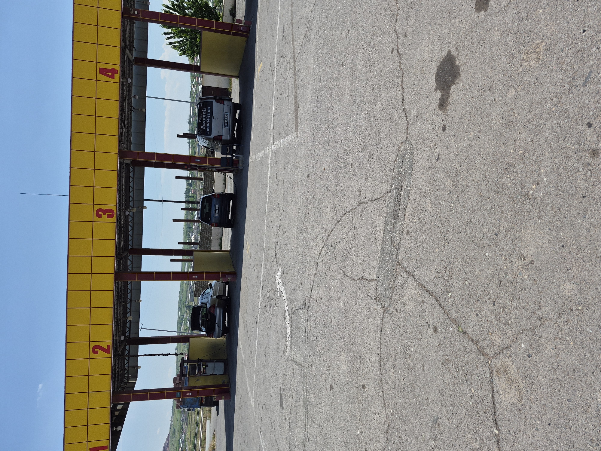 The image shows a view of a car wash facility in Ashtarak, Armenia. The facility has a yellow awning supported by red and yellow poles, with numbers 1, 2, 3, and 4 visible on the awning, indicating parking spots or bays. Several cars are present at the facility. In bay 2, a silver sedan with its trunk open is parked, and a gas pump is visible next to it, suggesting it is either being filled or has just been. In bay 3, a dark-colored SUV is parked. In bay 4, a silver minivan is parked, and its rear doors are open. The ground is paved with asphalt, showing signs of wear and cracking, with a few dark stains that appear to be oil or water. In the background, there are buildings, some vegetation, and hills under a clear blue sky. The overall scene depicts an outdoor car wash operation on a sunny day.