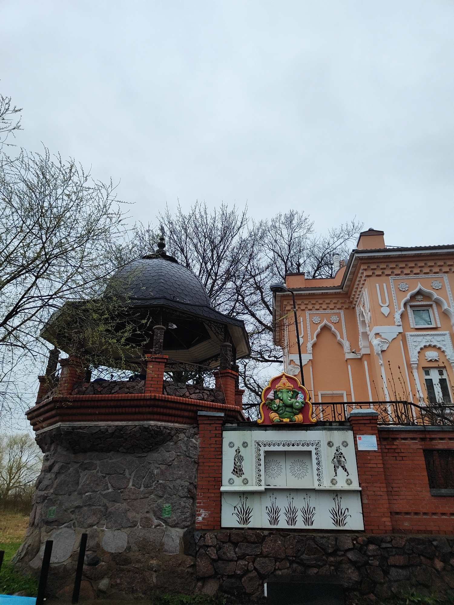 The image displays an architectural scene under an overcast, light gray sky, suggesting a cloudy day. On the left, a structure with a robust base of rough, dark-colored stones is topped by red brickwork. This brick section supports a dark, scaled dome, possibly made of metal or shingles, which is adorned with ornate metal railings. Bare tree branches with some emerging light green buds frame the dome.
To the right, a multi-story building features a distinct peach-colored facade with extensive white architectural ornamentation. This decoration includes arched window frames, intricate patterns, and traditional Indian motifs such as `U`-shaped symbols and floral designs. A black metal balcony railing extends from this building.
In the center foreground, a decorative boundary wall connects the two main structures. It has a base of red brick, above which are white panels decorated with black stenciled designs. A prominent, colorful statue of Ganesha, predominantly green with golden and red details, is centrally placed atop these white panels. The white panels feature a depiction of a woman in traditional attire on the left, a stylized decorative border, and a male figure, possibly a warrior, on the right. Below these, black stylized plant motifs are visible. A small, blue sign with an indiscernible logo and text is affixed to the red brick wall section to the right of the white panels. No human activity or people are visible in the scene.