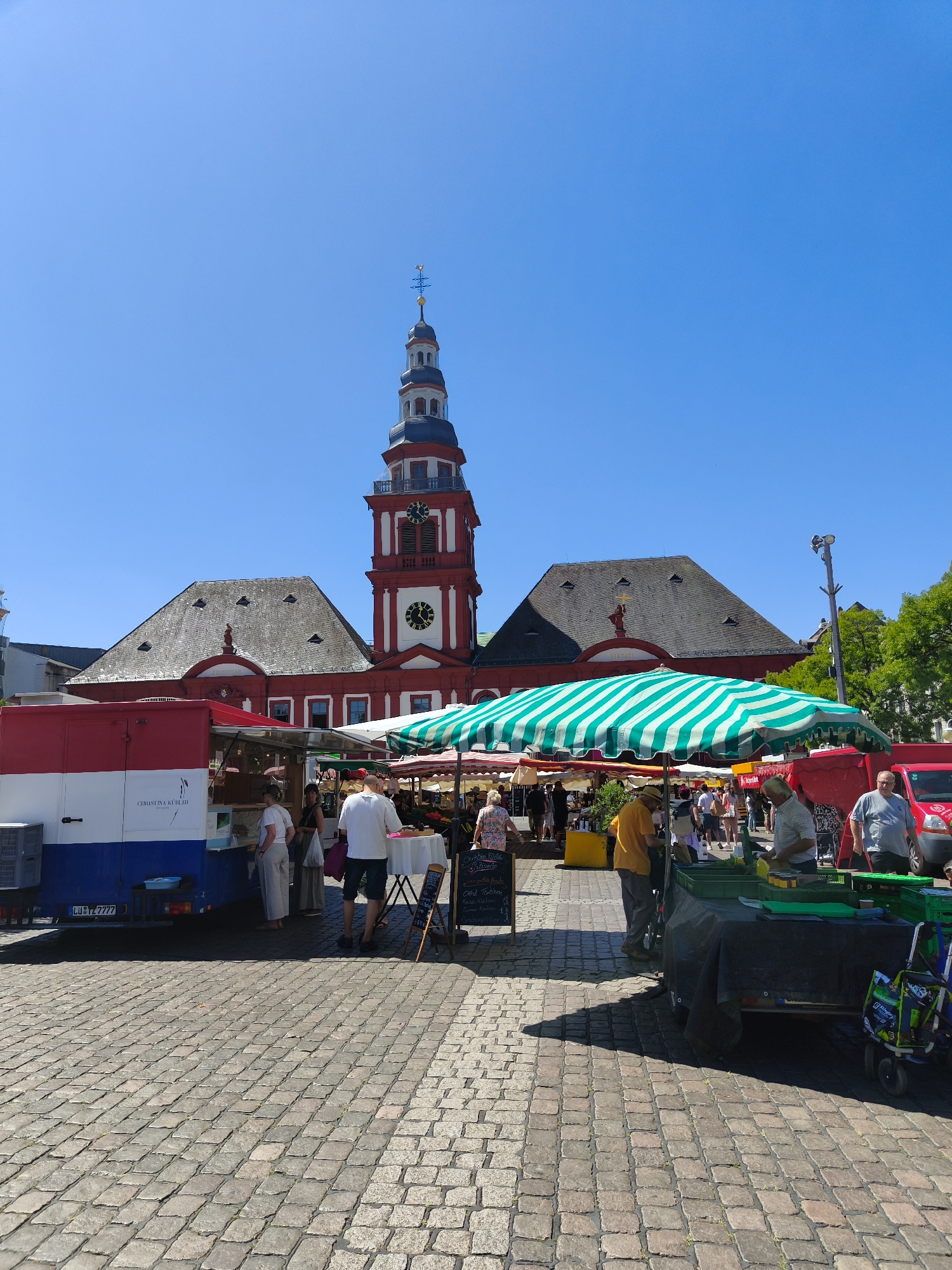 This is a full, eye-level, outdoor shot of a market square in Mannheim, Germany, on a clear, sunny day with a bright blue sky. In the background, a red and white building with a prominent clock tower stands tall. The clock tower has a white face with black Roman numerals and a spire. To the left of the clock tower is a large, dark-tiled roof with visible skylights.
In the foreground, a cobblestone plaza is bustling with activity. Several market stalls are set up. One stall, to the left, is a red, white, and blue food truck with text reading "CHRISTINA KÜCHLER". Further to the right, a large, green and white striped umbrella shades a produce stall. A variety of people are visible browsing and interacting at the stalls.
There are at least five visible market stalls. The people appear to be a mix of vendors and shoppers, dressed in casual summer clothing. A red vehicle is parked to the far right of the frame. The scene depicts a vibrant, daytime market atmosphere in a European city.