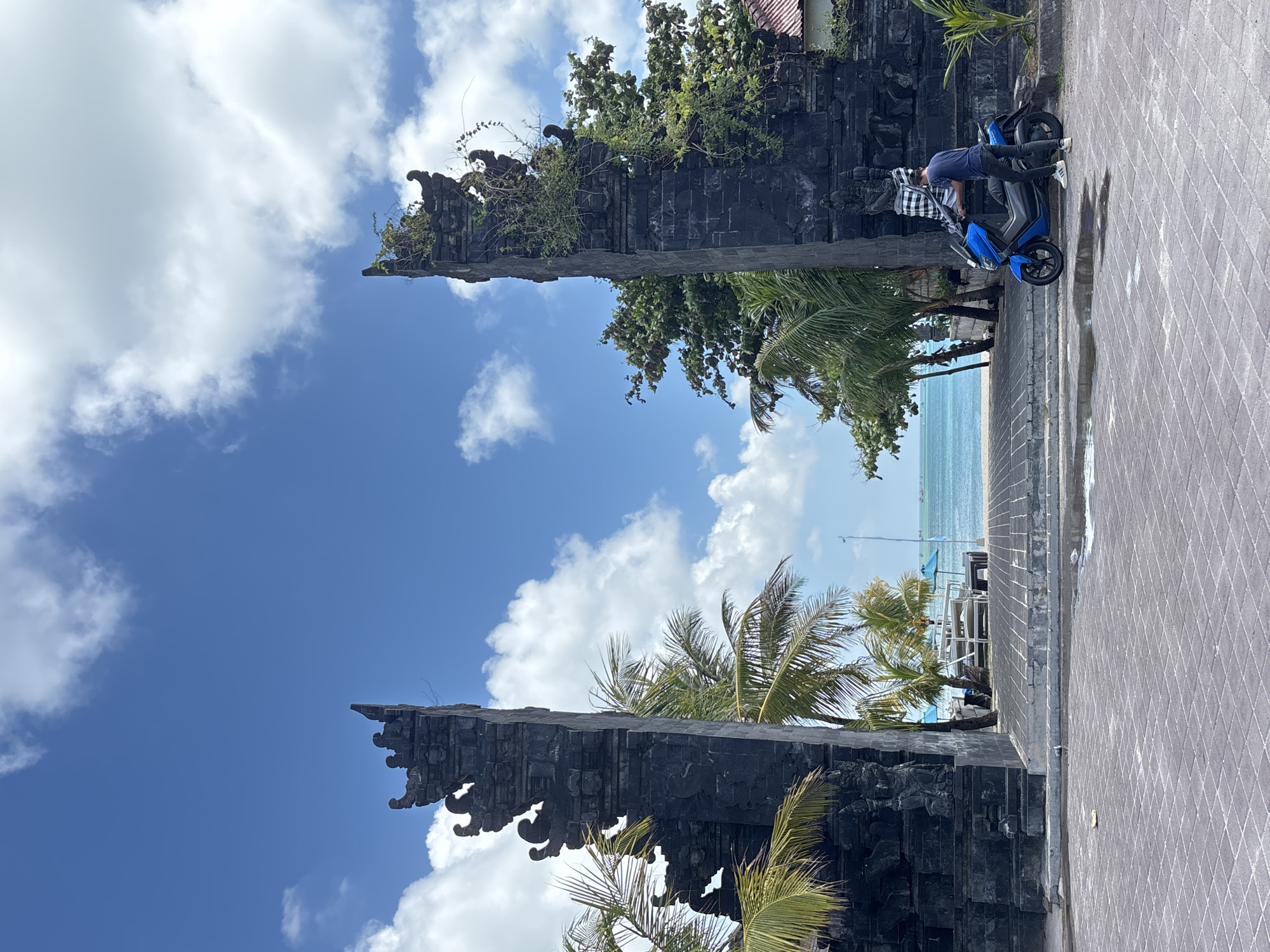 A vertical shot shows the iconic gate of Kuta, Indonesia, framed by a blue sky dotted with fluffy white clouds and tropical foliage. The imposing, black stone structure is a key landmark, with intricate carvings. A person is crouched near a blue scooter, their attention seemingly directed toward the vehicle. The background reveals a glimpse of the ocean and beach, furthering the scene's coastal context. The foreground presents a paved surface, indicating the location is a public area, likely a pathway or sidewalk.