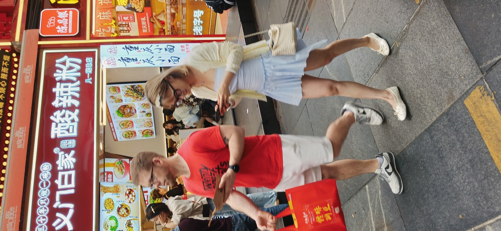 The image captures a street scene in Yuzhong District, China, featuring two pedestrians passing in front of a food stall or restaurant. A man wearing a red t-shirt and white shorts walks on the right-hand side, holding a red shopping bag and looking at his phone. Beside him, a woman in a light blue dress and cardigan is also looking at her phone. Both of them are walking on a paved sidewalk next to the restaurant. The restaurant is lined with food advertisements and Chinese text, indicating the offerings of a local establishment. Behind the pedestrians, through the windows of the restaurant, other people can be seen sitting and eating at tables. The scene is captured during the daytime.